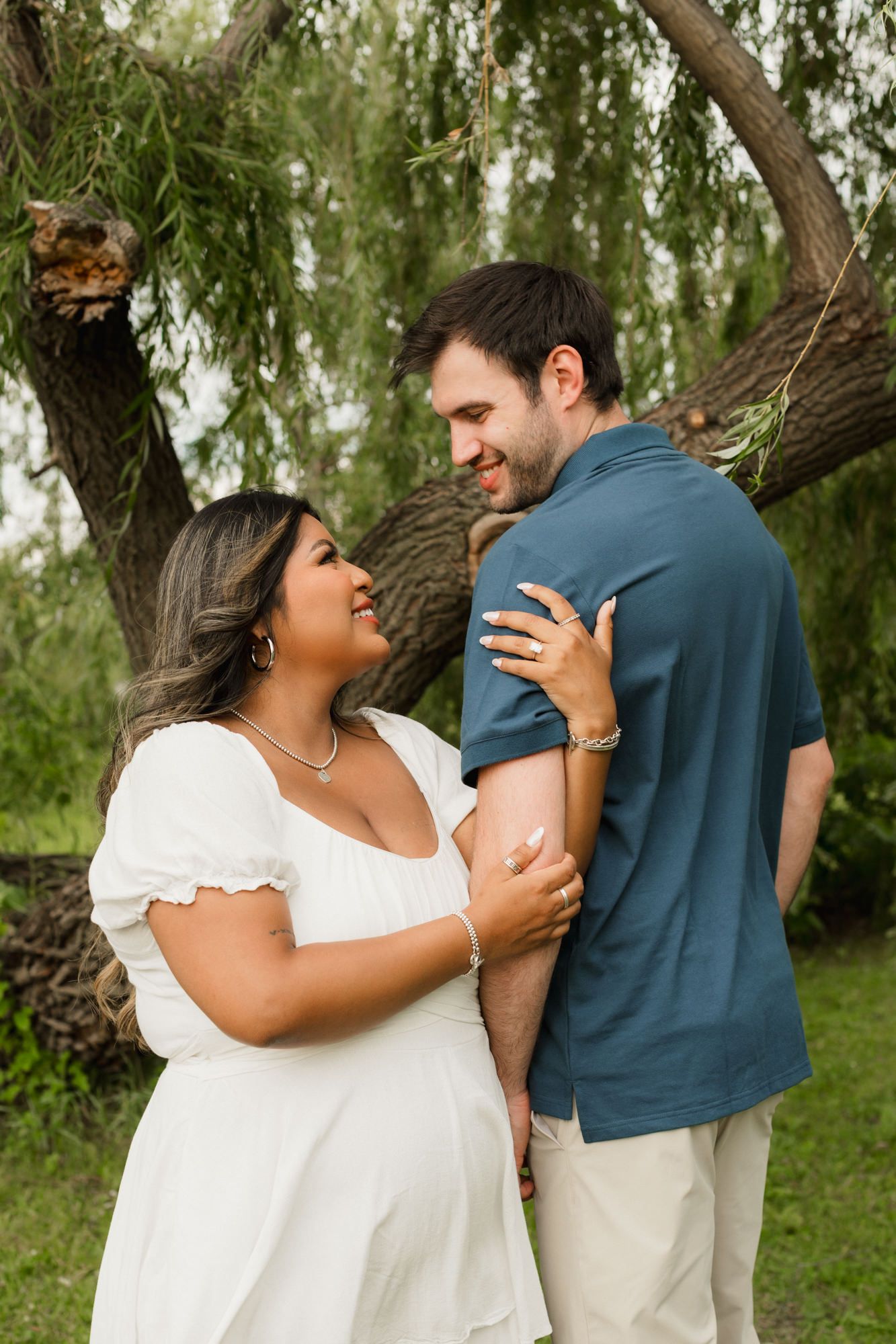 A couple shares a tender moment outdoors near a willow tree, woman in white dress touching man's arm.