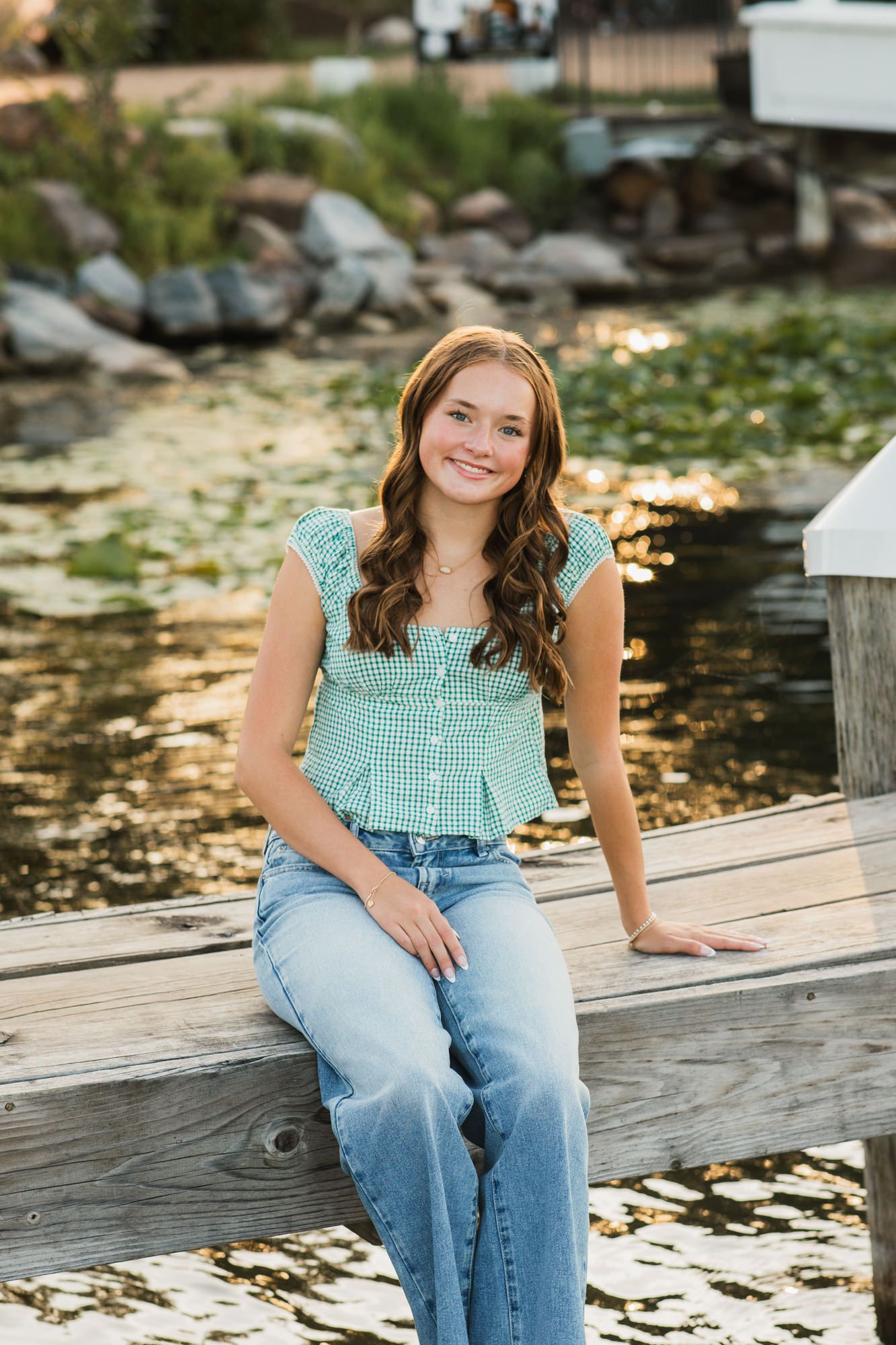 Young woman in green gingham top and jeans sitting on a wooden dock by a serene pond, smiling warmly.
