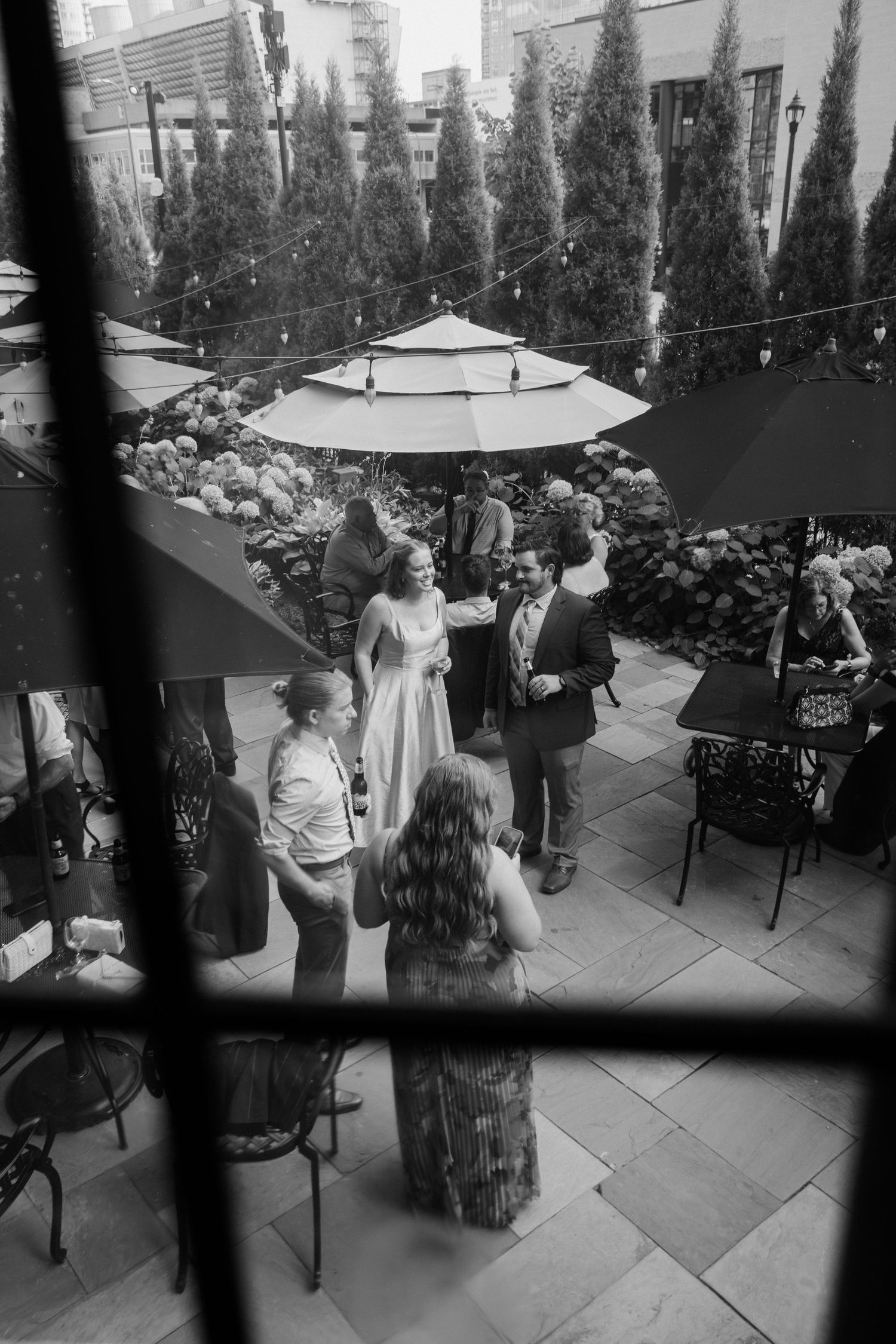Black and white aerial view of elegantly dressed guests mingling on a patio with umbrellas and cypress trees.