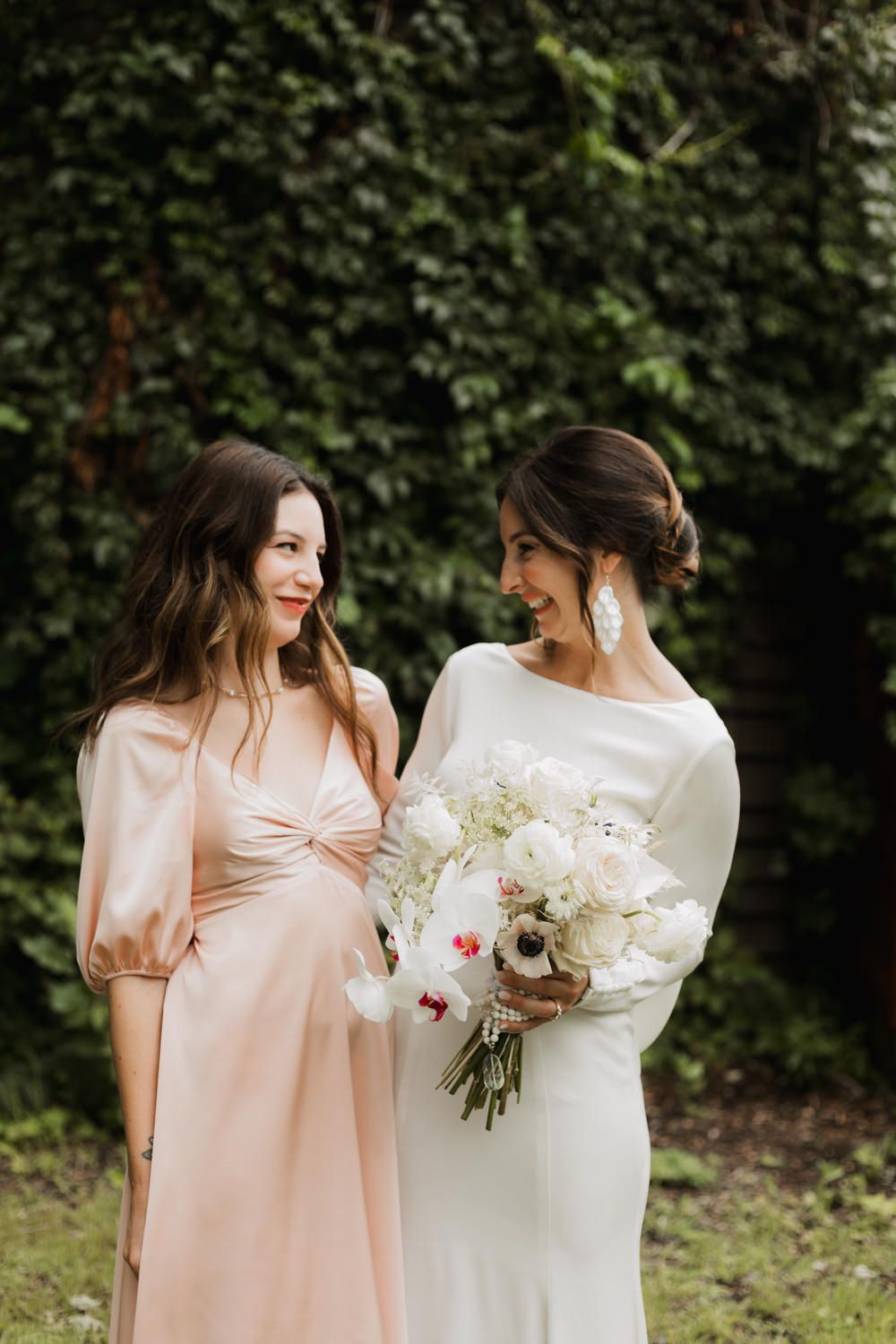Two women in white and blush dresses share a joyful moment against a green ivy backdrop holding a white floral bouquet.