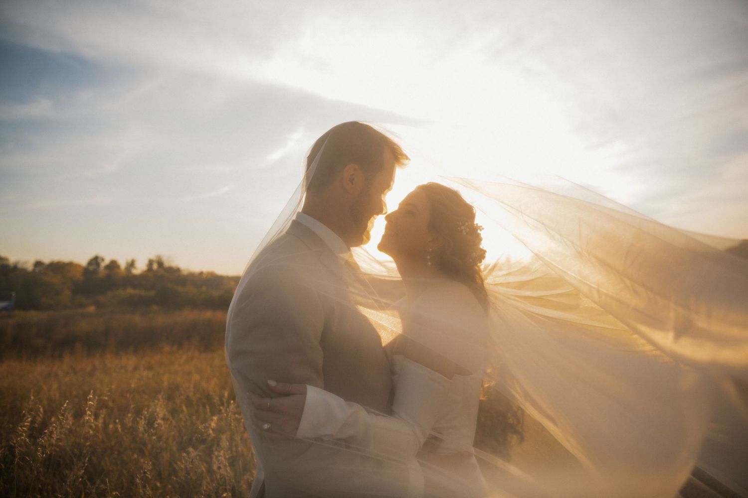 Bride and groom embrace in a sunlit field as her sheer veil blows around them in warm golden light.