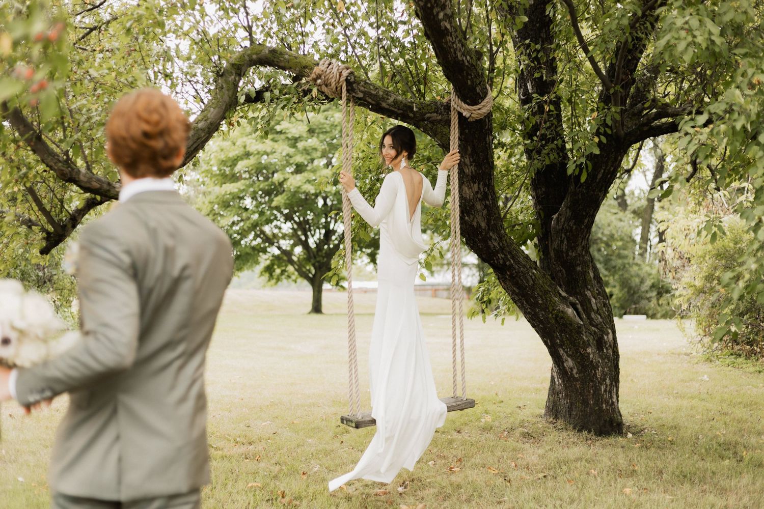 Bride in white gown standing on a swing tied to a tree, with groom in gray suit watching in a garden setting.