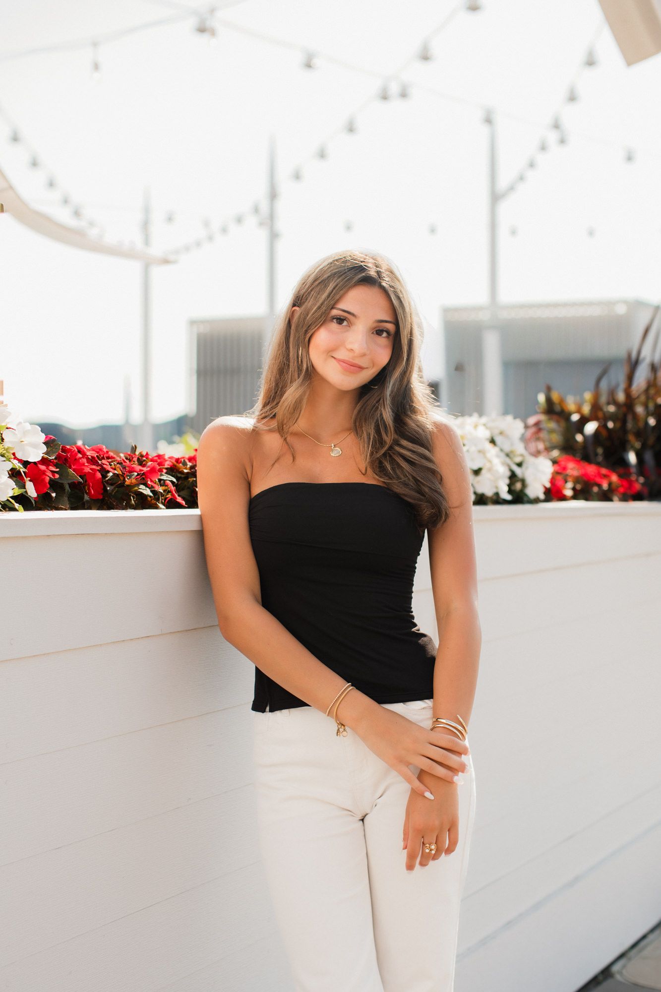 Young woman in black tube top and white jeans leaning against white wall with red and white flowers.