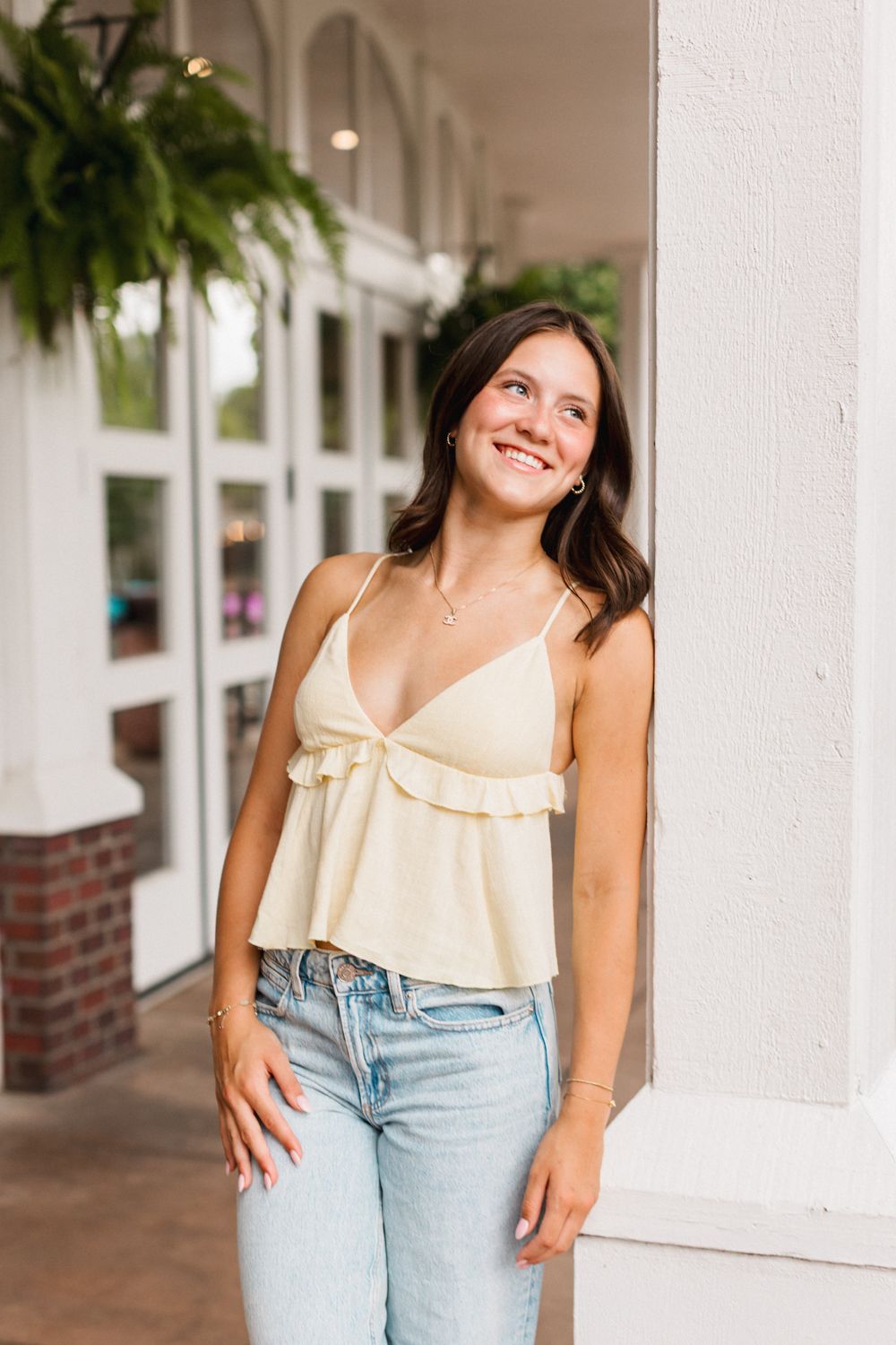 Young woman in cream colored tank top and light jeans smiles while leaning against white column on covered porch.