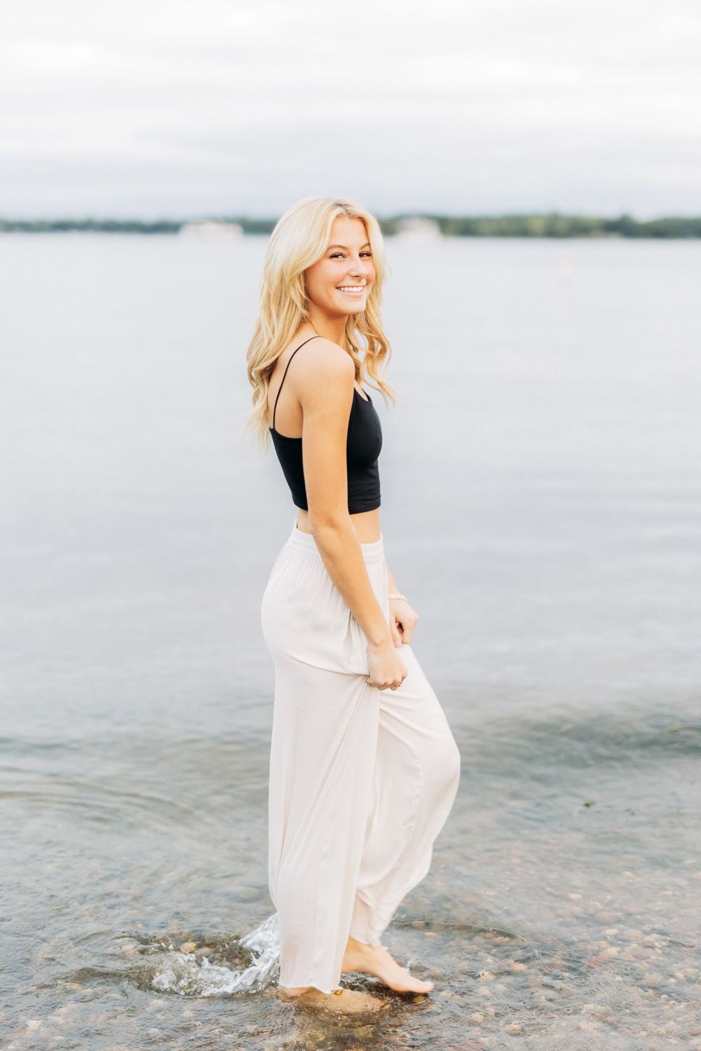 Woman in black top and white skirt splashes and plays joyfully at the water's edge on a cloudy day.
