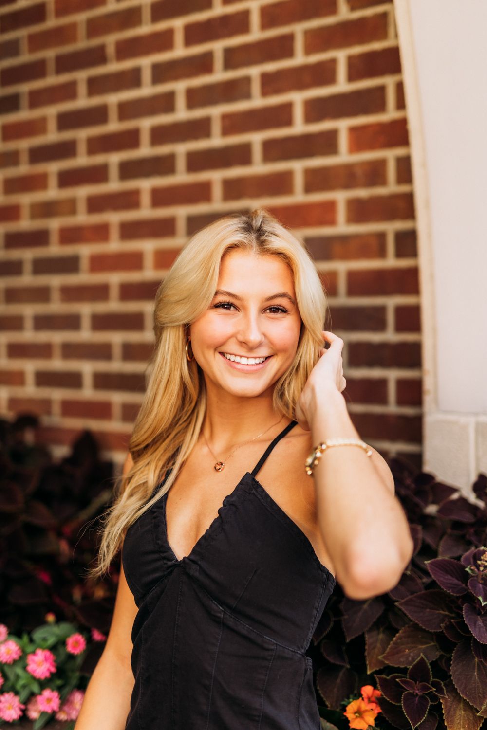 Woman in black sleeveless dress smiles while posing against brick wall with pink flowers.
