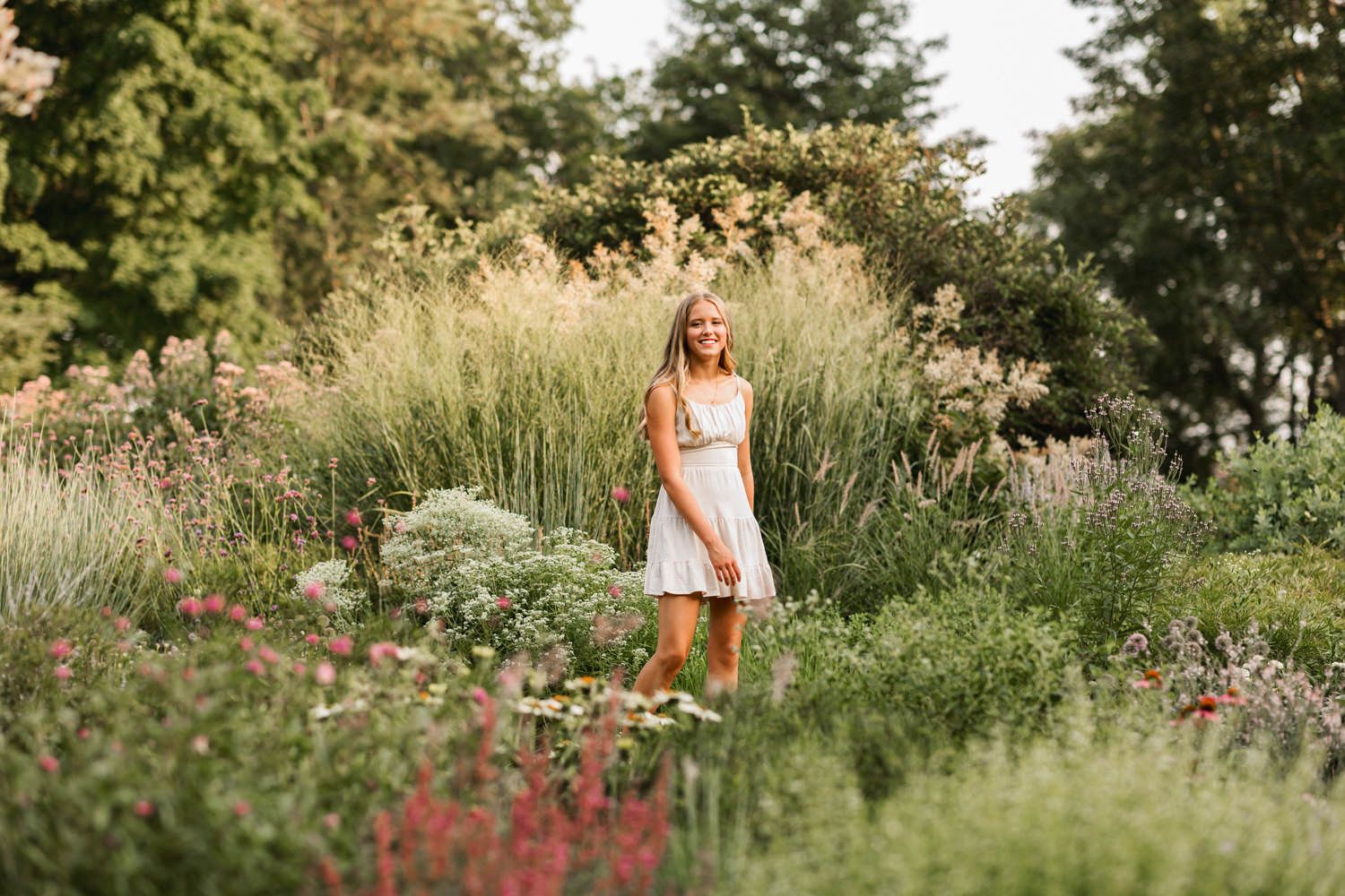 A figure in a white dress stands amid a lush summer garden filled with tall grasses and wildflowers at sunset.