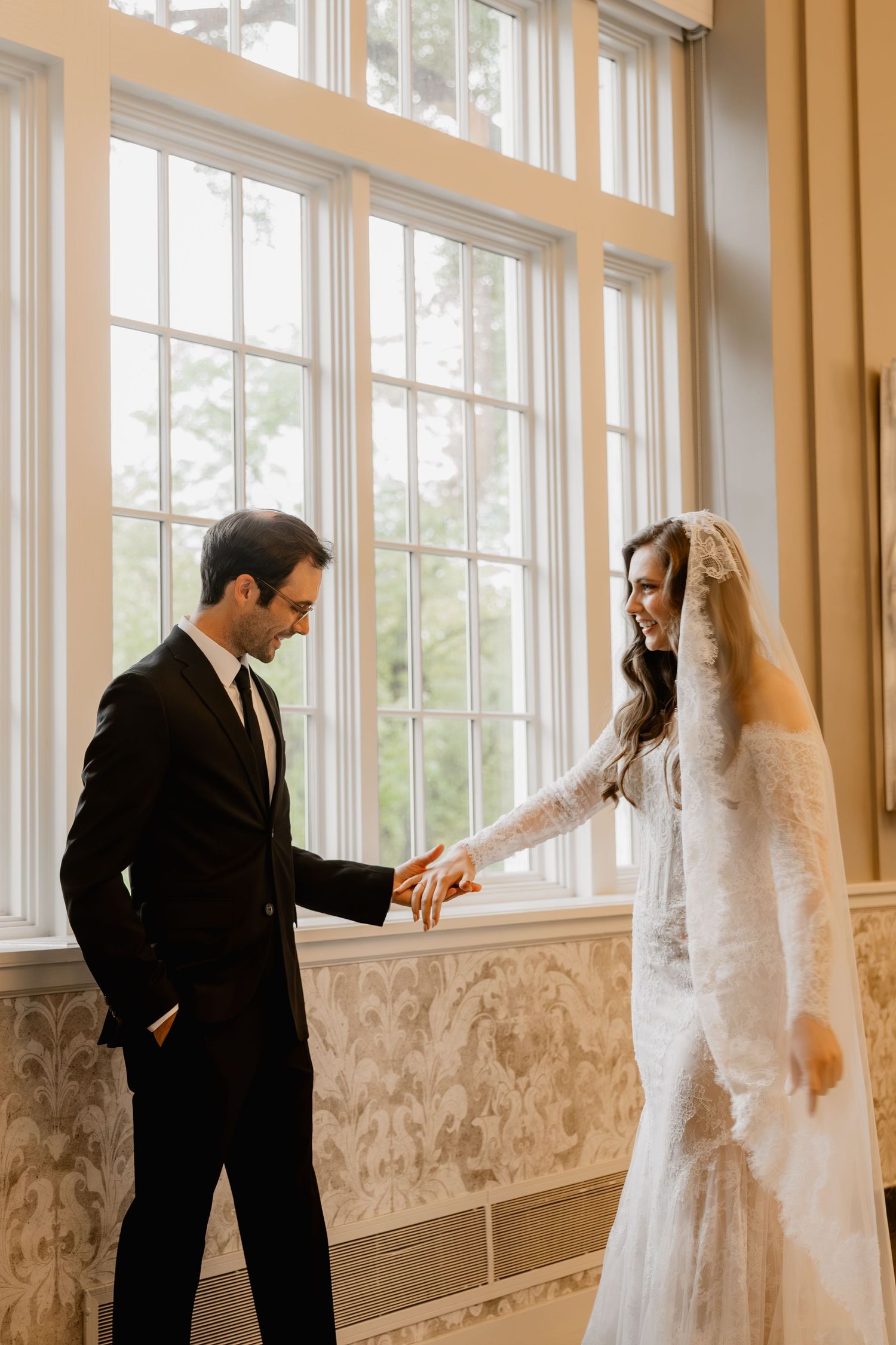 A groom in a black suit holds his bride's hand as she smiles in a lace wedding gown and veil by large windows.