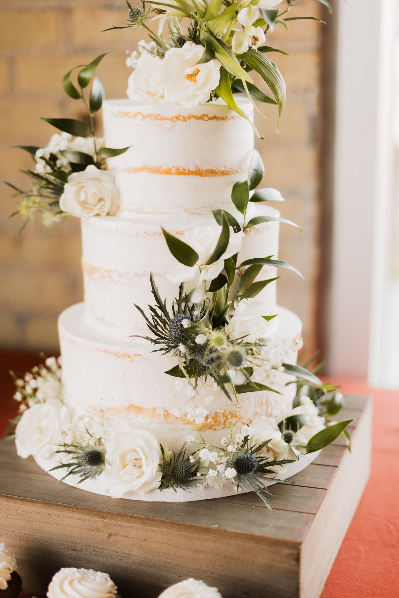 Four-tier semi-naked wedding cake adorned with white roses, greenery, and thistle flowers on a rustic wooden stand.