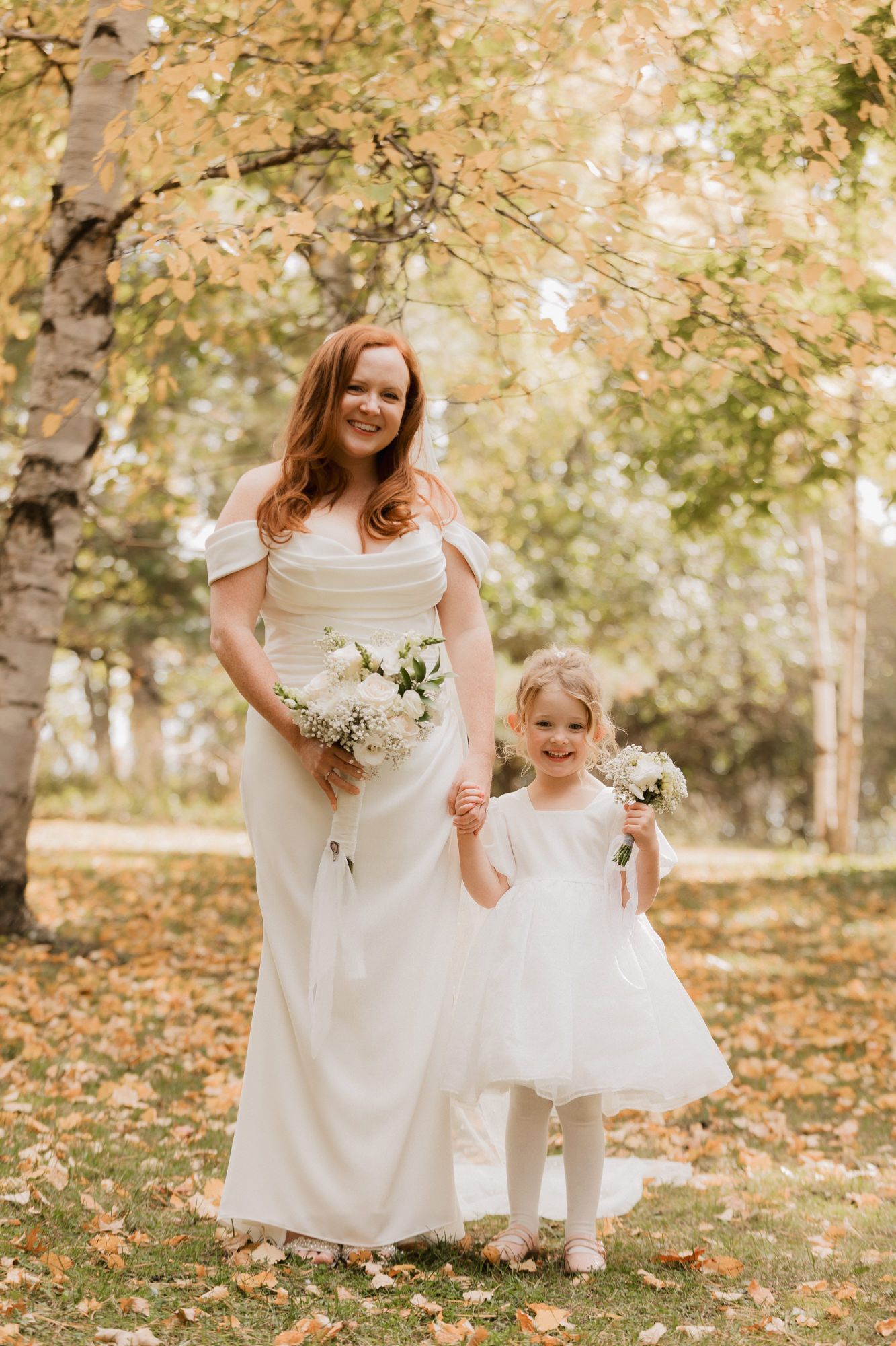 Bride in white gown holds hands with flower girl in autumn park, both carrying white bouquets.