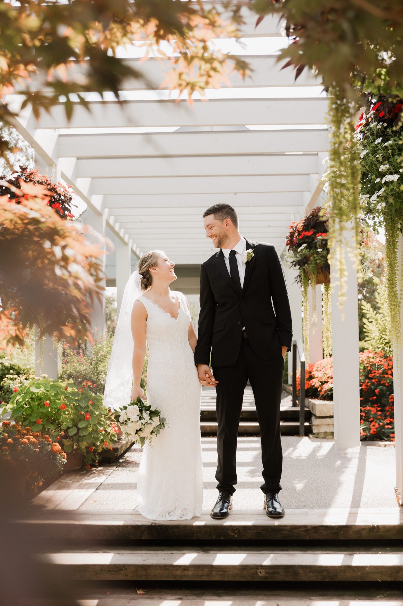 Bride and groom holding hands smiling at each other under a white pergola surrounded by colorful garden flowers.