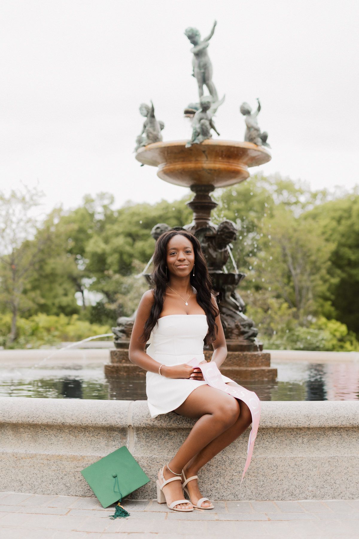 Young woman in white dress sits by ornate fountain with green graduation cap, celebrating her achievement outdoors.