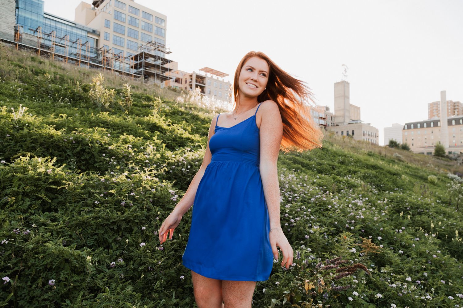 Young woman with red hair in blue dress smiling outdoors with green hillside and city buildings behind her.