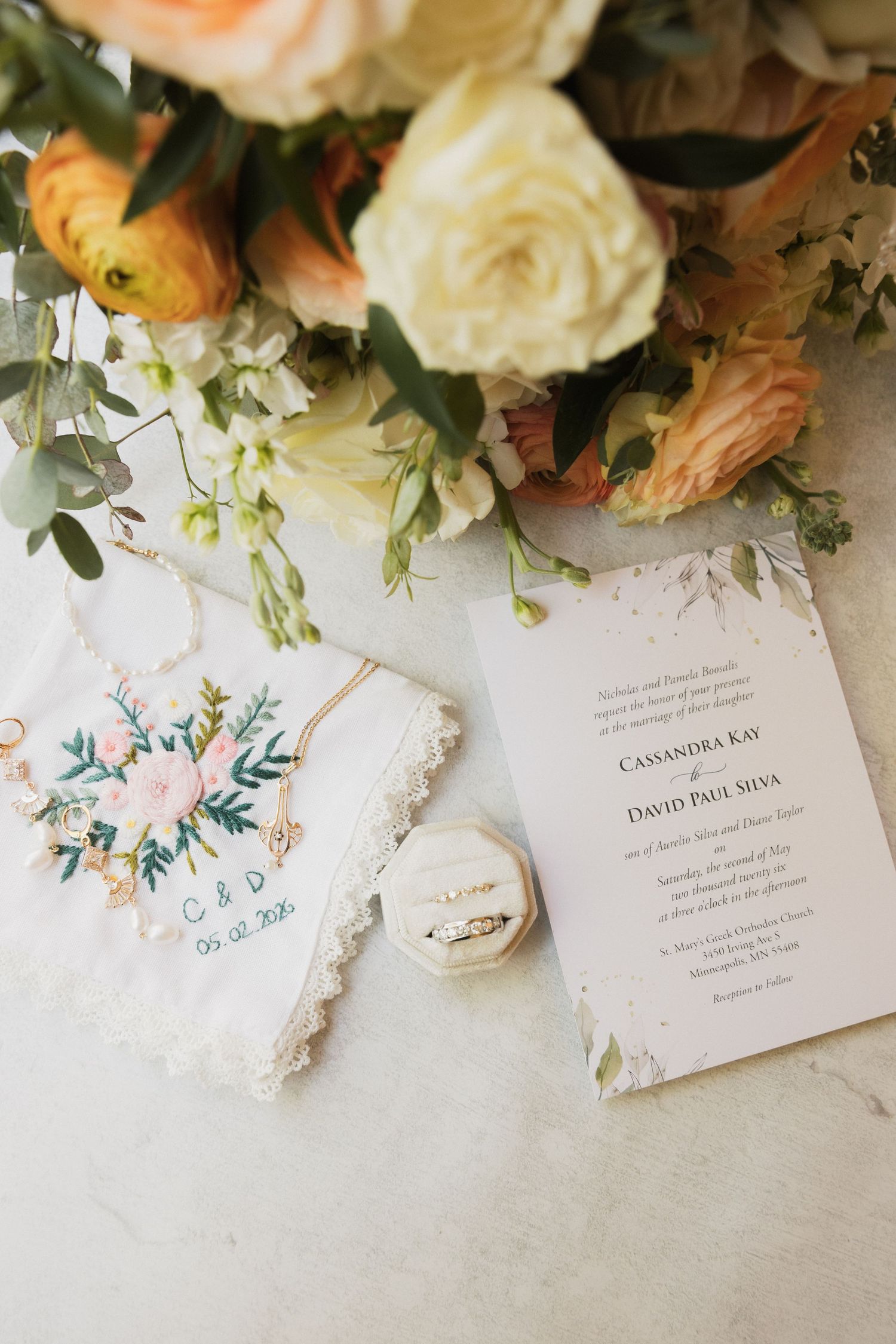 Wedding flat lay with floral bouquet, embroidered handkerchief, ring box, and elegant invitation for Cassandra and David.