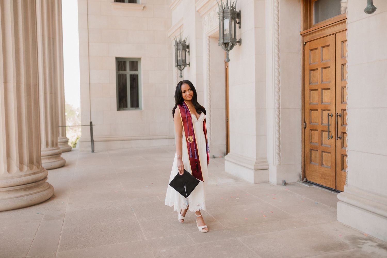 A smiling graduate in white dress and maroon stole holds cap outside a grand stone building with columns.