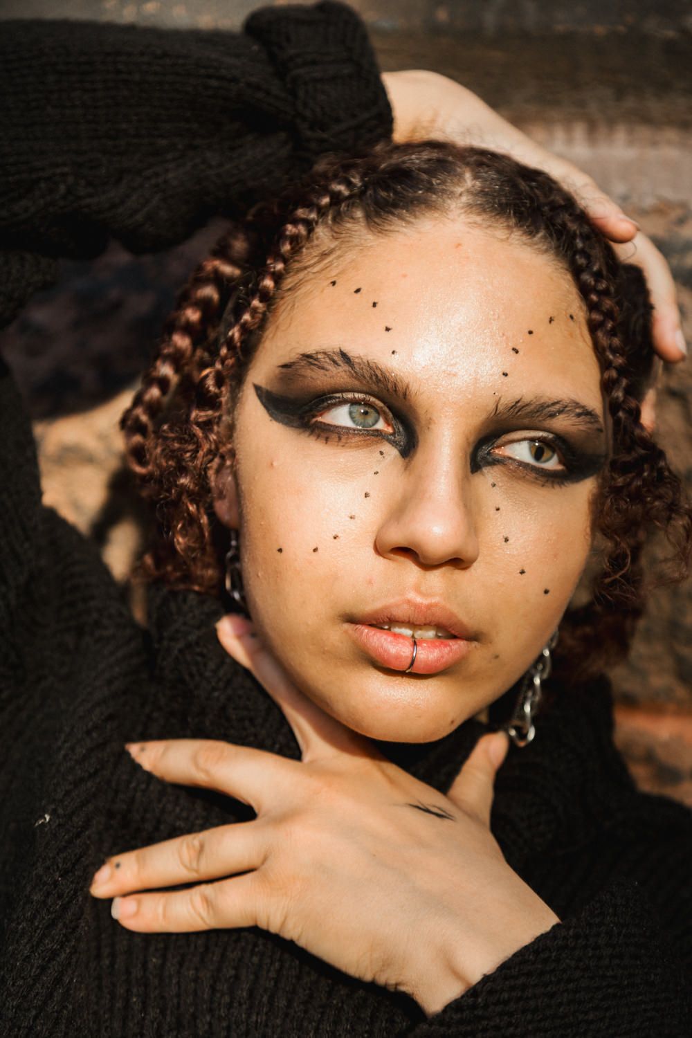 Young woman with dramatic black smoky eye makeup, dot freckles, braided hair, wearing a black turtleneck sweater.