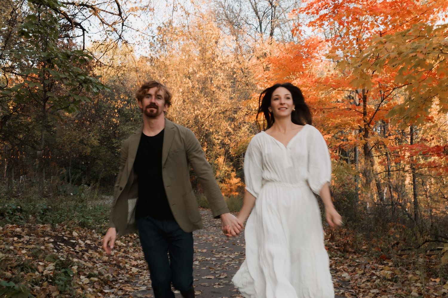 Couple holding hands walking through autumn forest with vibrant red and orange foliage, woman in white dress.