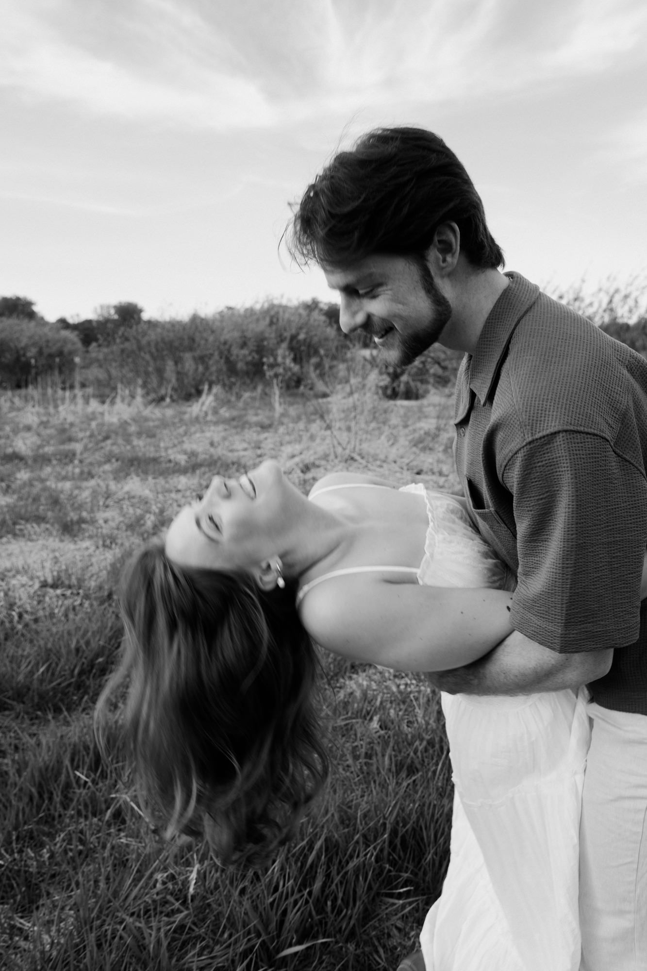 Black and white photo of a smiling couple in a field, man holding woman as she leans back laughing.