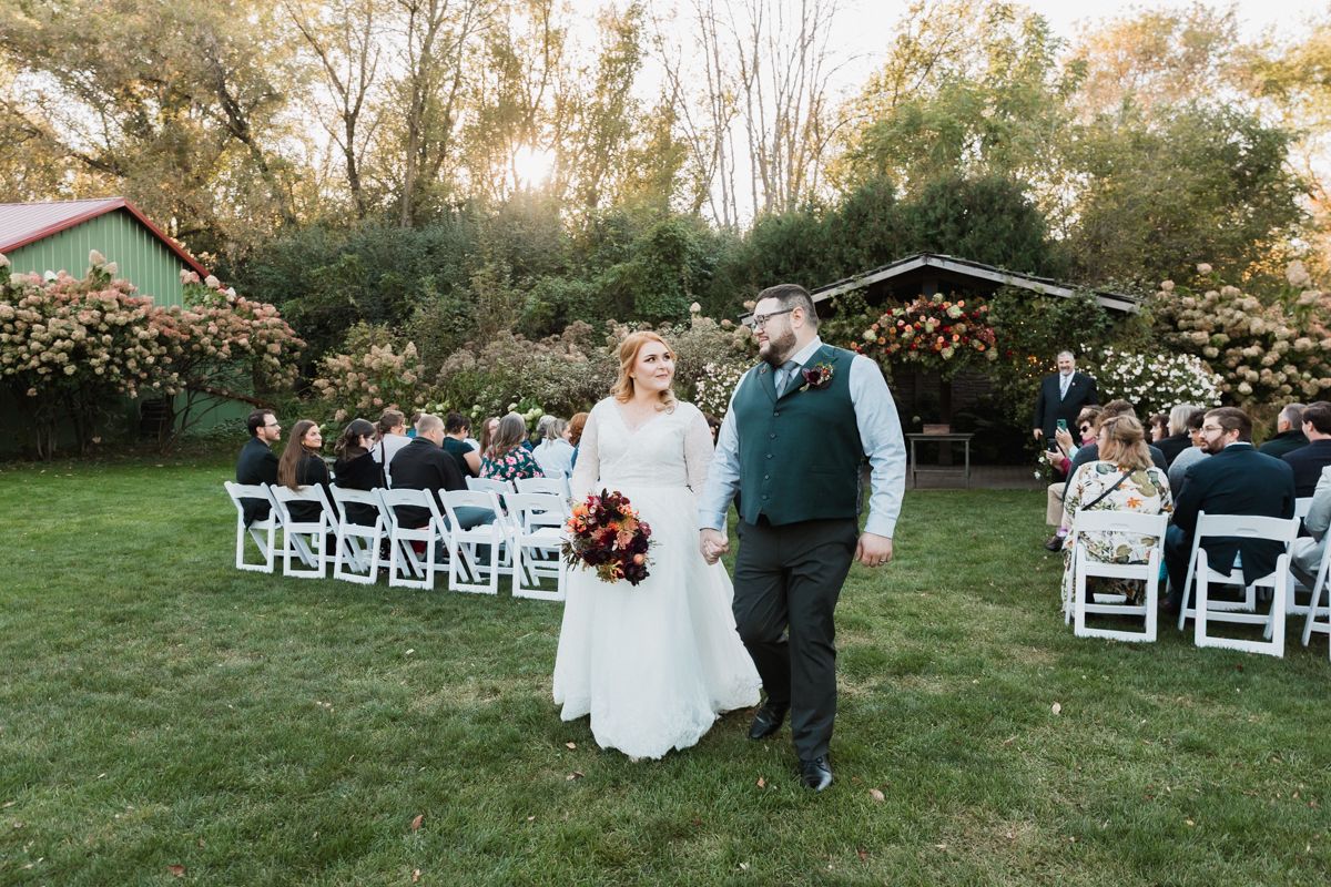 Wedding ceremony in progress in a lush garden setting with white chairs and autumn foliage.