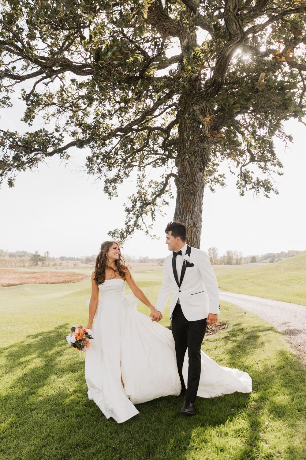 A romantic wedding couple dances together under a sunlit tree on a grassy hillside during golden hour.