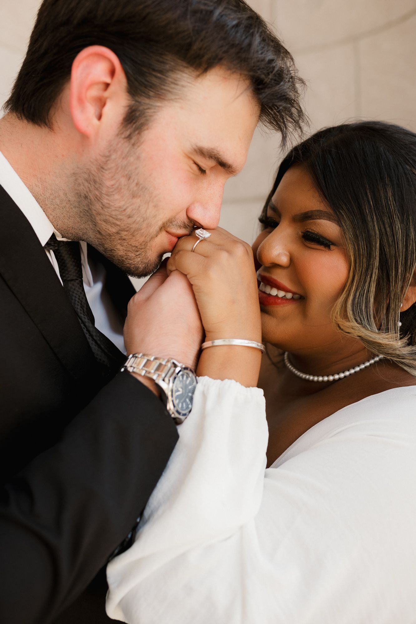 A groom in a black suit kisses his bride's hand as she smiles, wearing a white dress and pearl necklace.