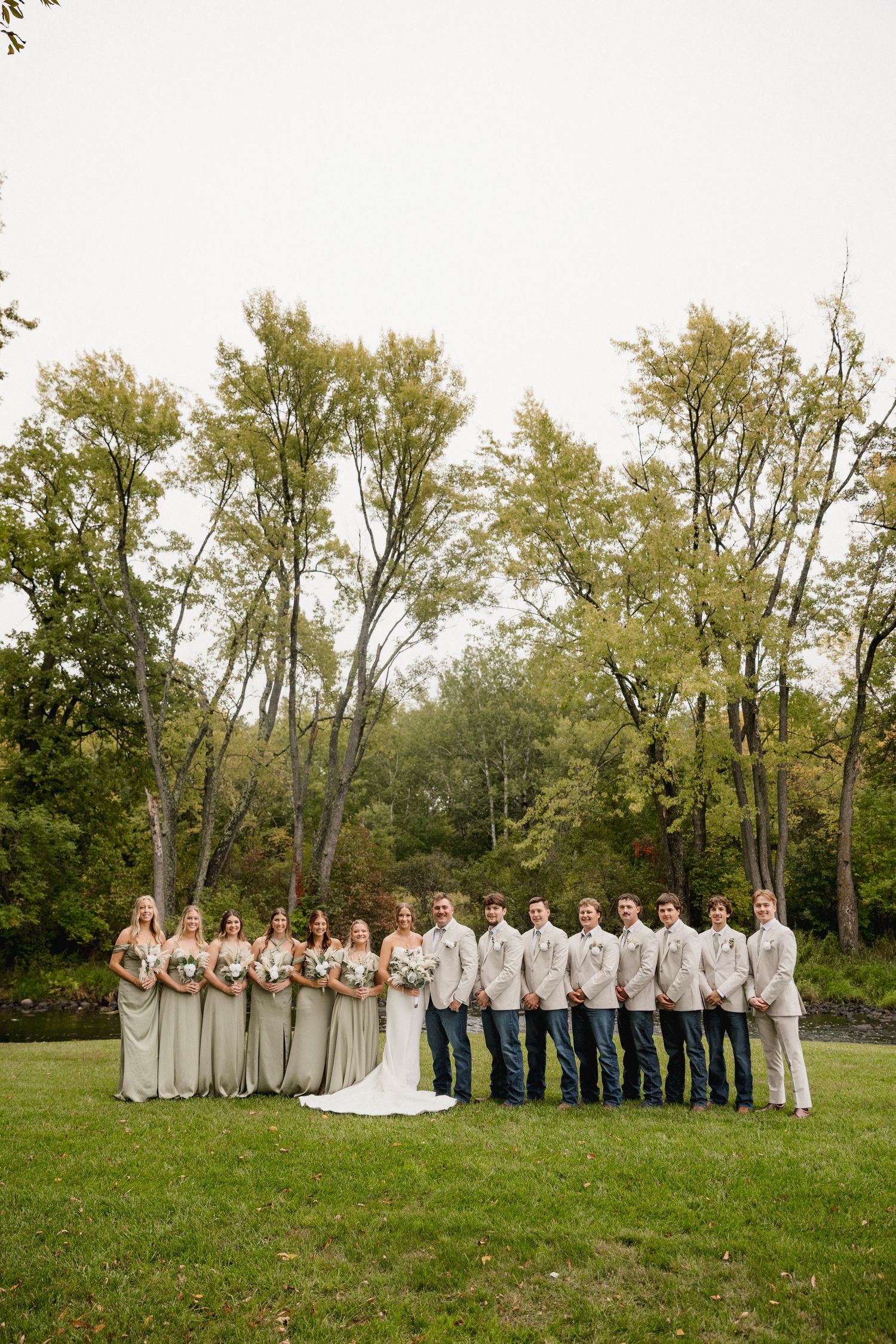 Wedding party in neutral tones poses outdoors on green lawn with lush trees, bridesmaids in sage gowns and groomsmen in jeans.
