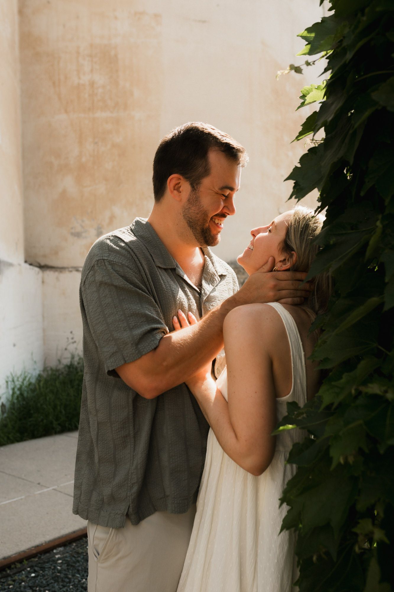 A couple shares a romantic moment outdoors, the man gently holding the smiling woman's face near lush green vines.