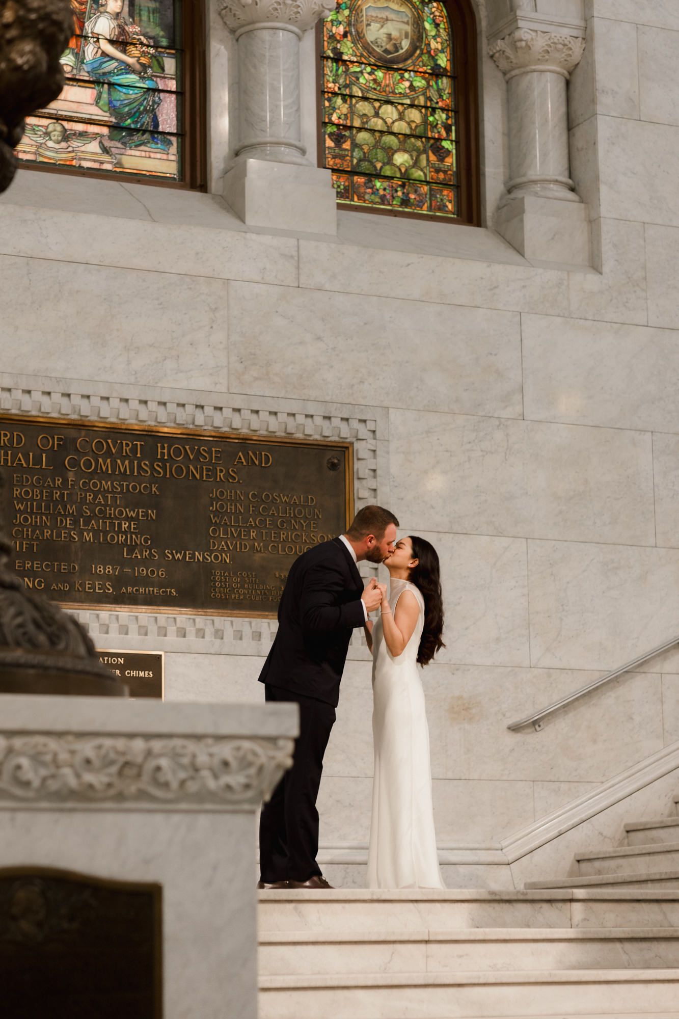 Bride and groom sharing a kiss on marble steps inside an elegant courthouse with stained glass windows.