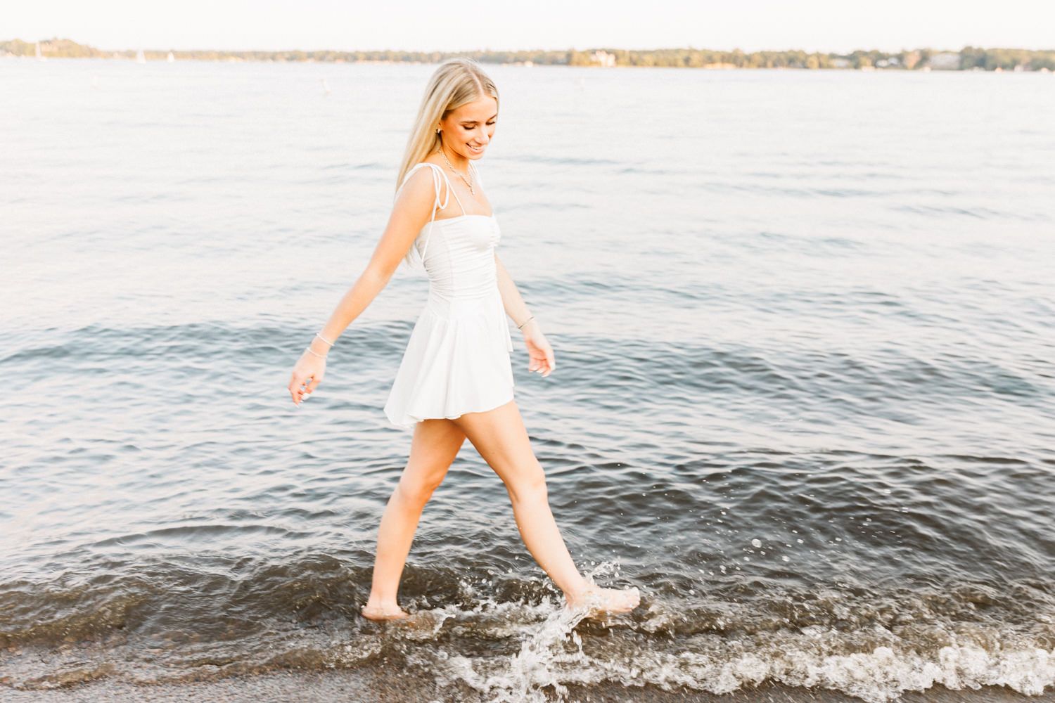 Person in white dress walking through shallow water at beach during sunset.