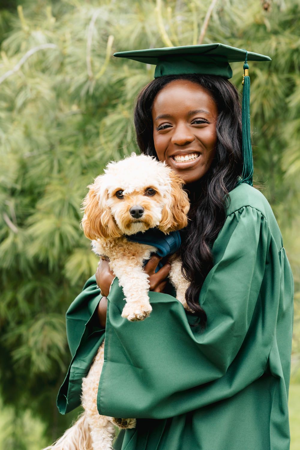 Graduate in green regalia holds small cream-colored dog wearing matching bow tie.