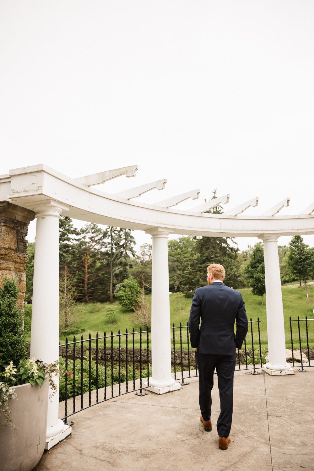 A groom in a black suit walks towards a white garden pergola on a cloudy day.