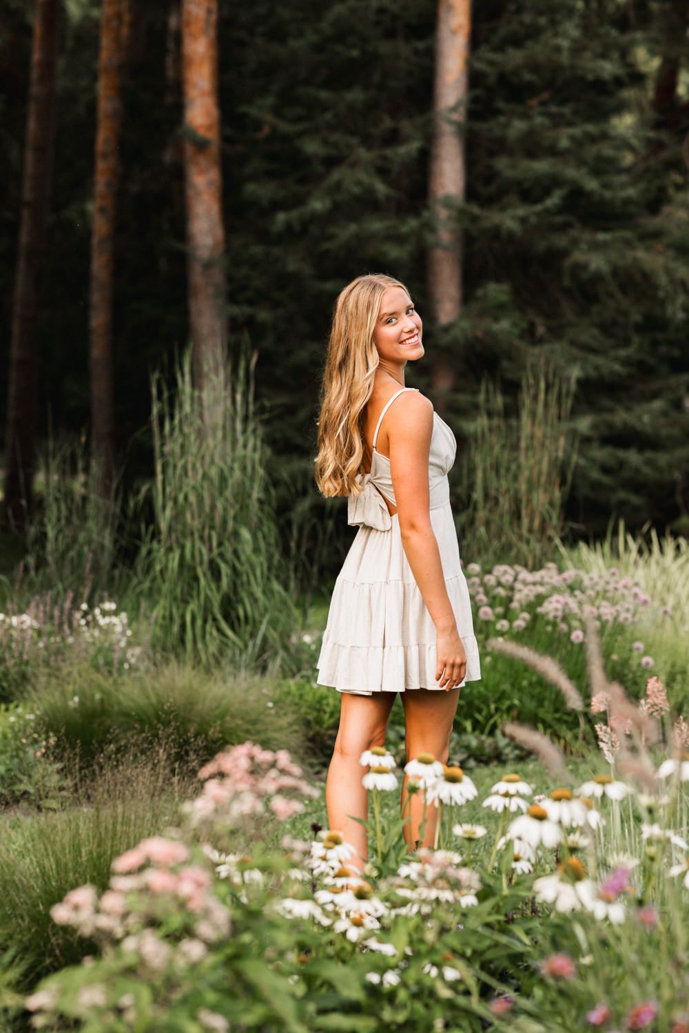 Someone wearing white dress poses in garden with daisies and pine trees in background.