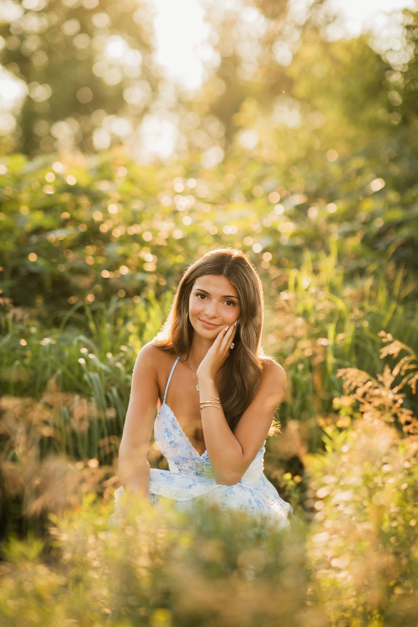 Young woman in a floral sundress sitting in a sunlit garden, smiling softly with golden bokeh background.