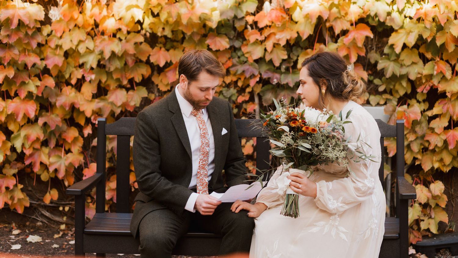 Bride and groom sitting on a bench reading vows, surrounded by colorful autumn foliage backdrop.