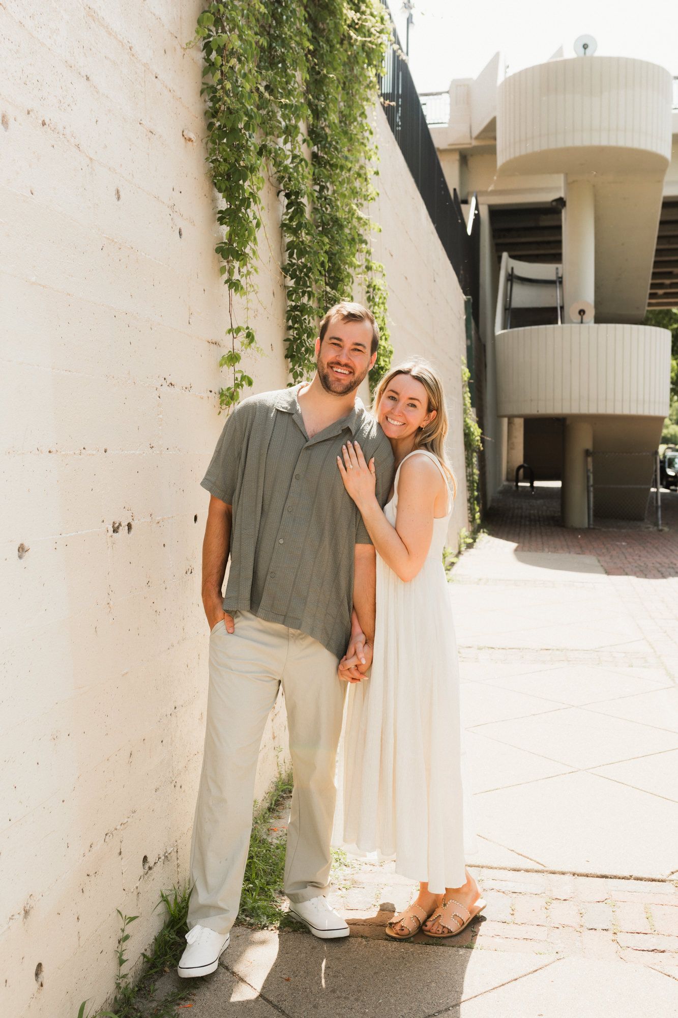 A smiling couple poses against a white brick wall with ivy, man in gray shirt and woman in white dress holding hands.