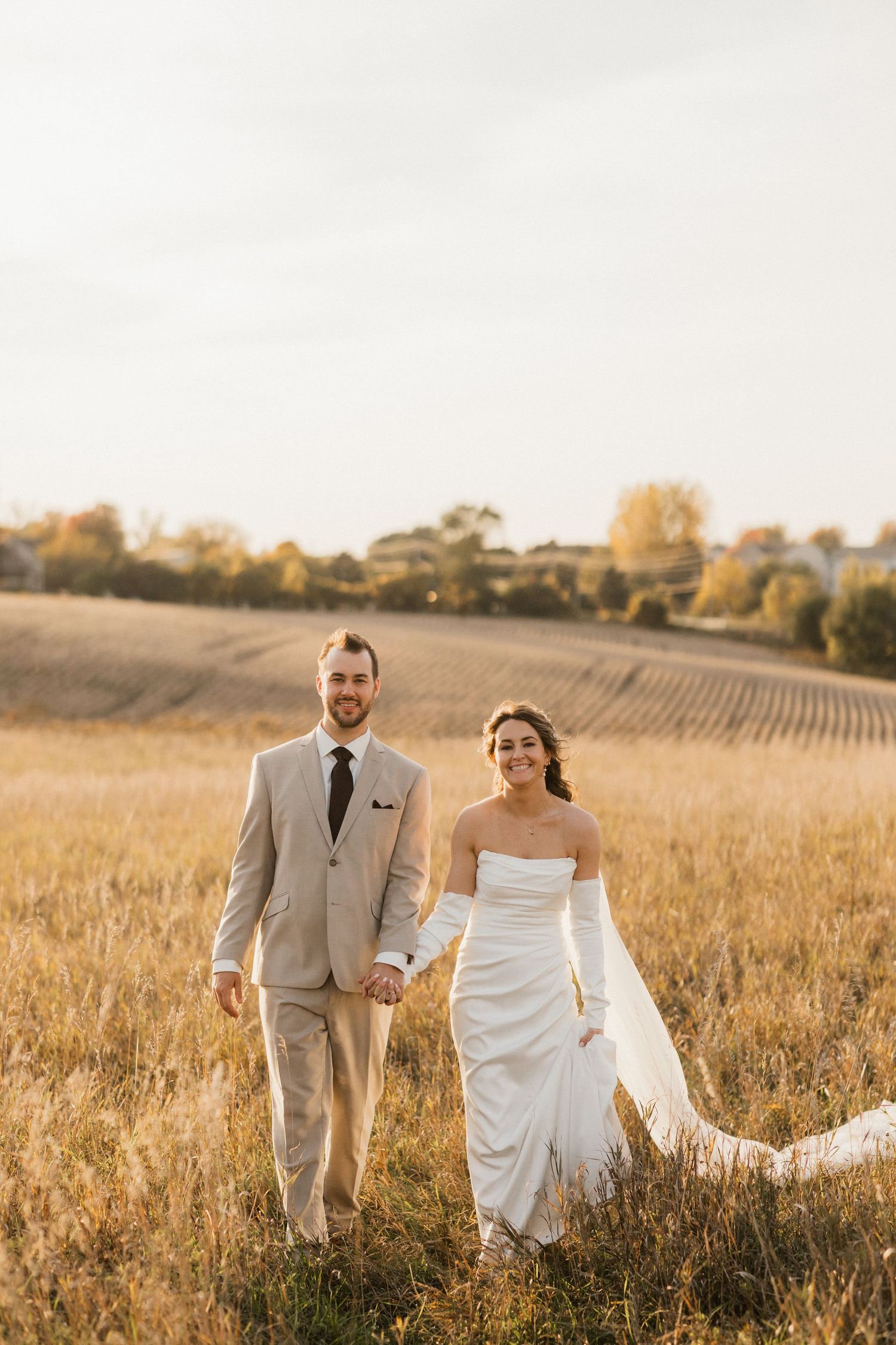 Bride and groom hold hands while walking through a golden field at sunset, her veil trailing behind.