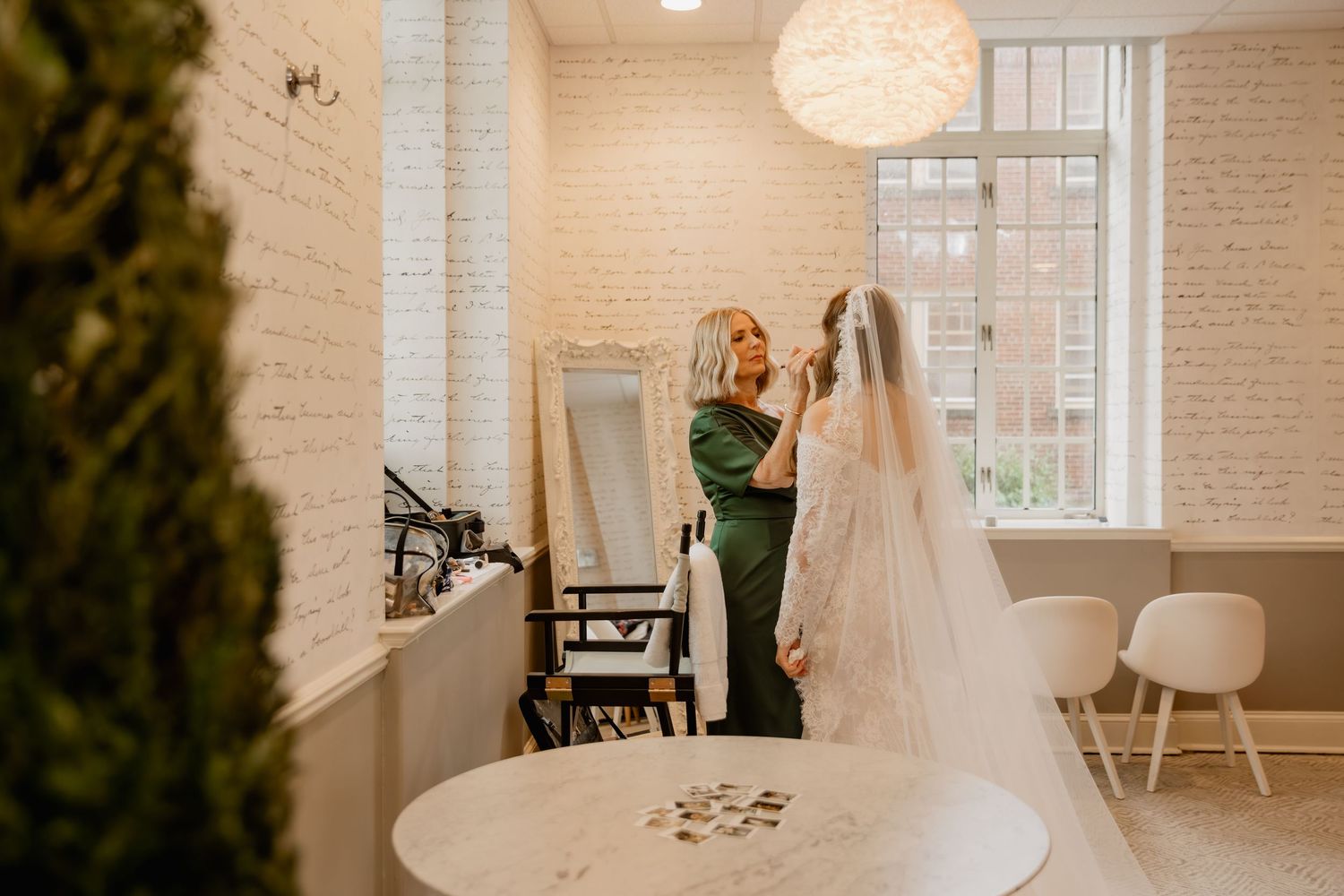 Bridesmaid in green dress adjusting bride's veil in bright white brick bridal suite with mirror.