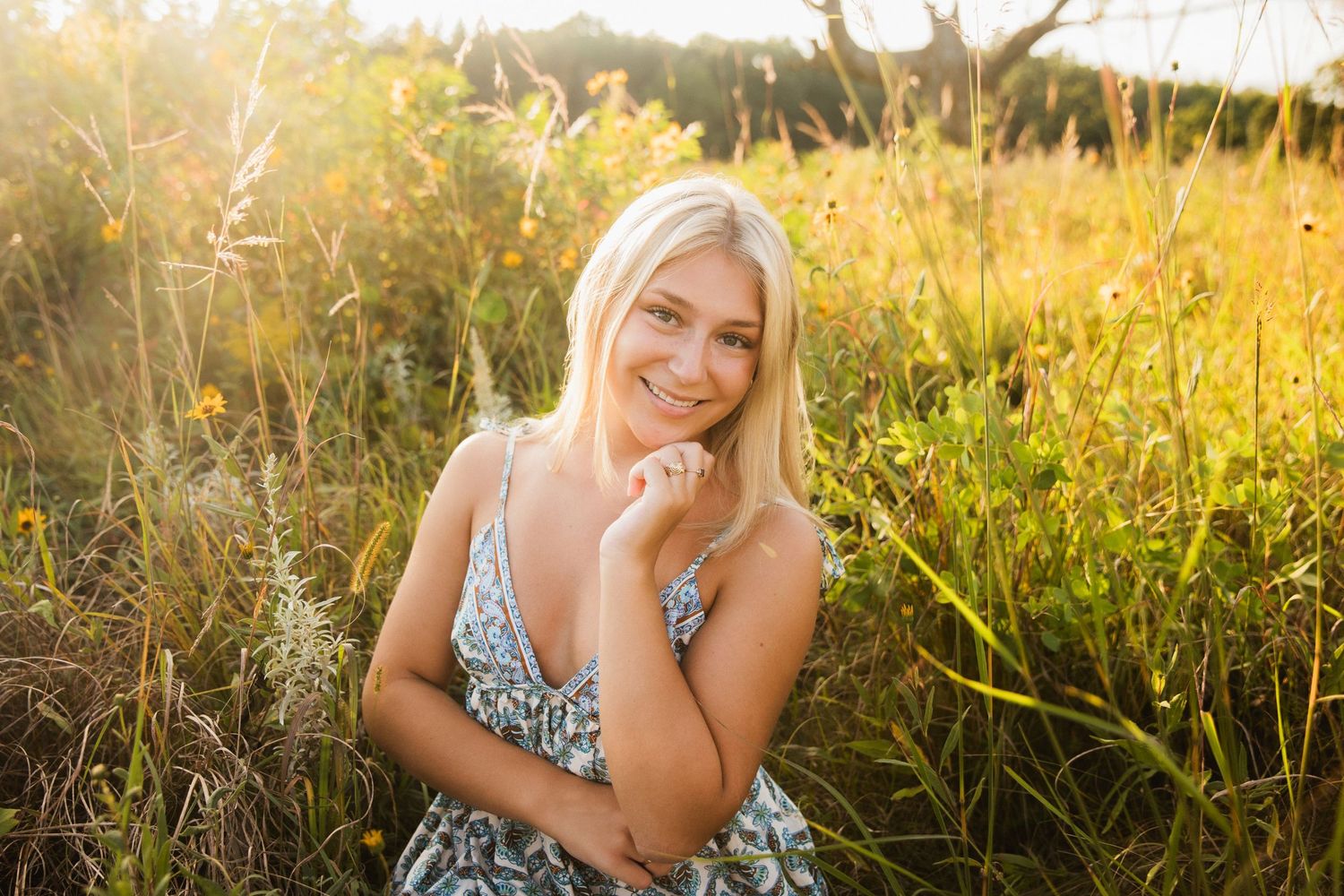 Smiling blonde woman in floral dress sitting in golden sunlit meadow with wildflowers during sunset.