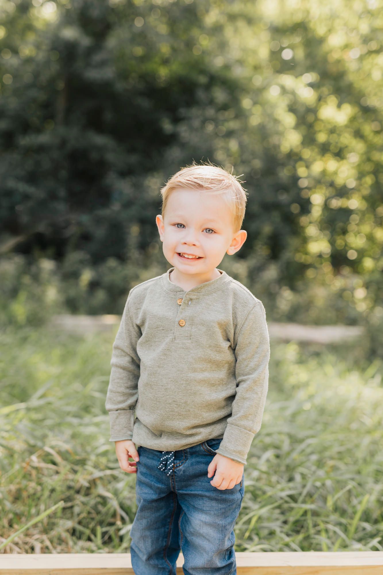 Toddler in gray sweater and jeans smiles while playing outdoors in natural setting.