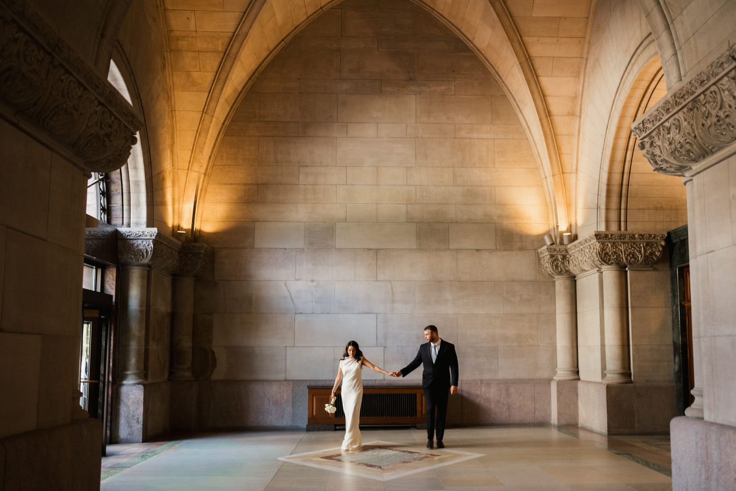 Couple holds hands in grand stone archway hall with warm golden lighting during elegant wedding ceremony.