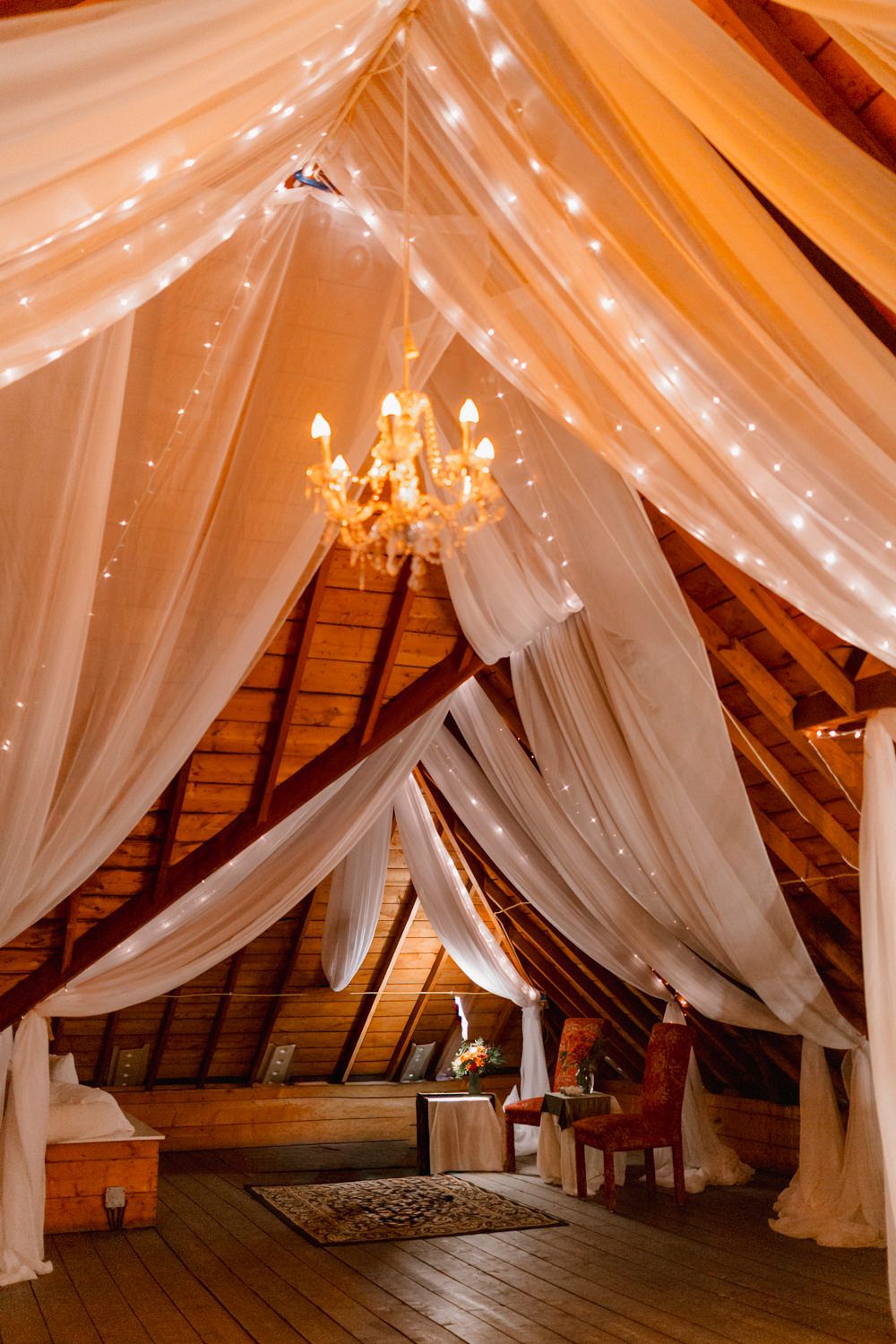 Romantic attic space decorated with flowing white fabric drapes, twinkling lights and an elegant chandelier.