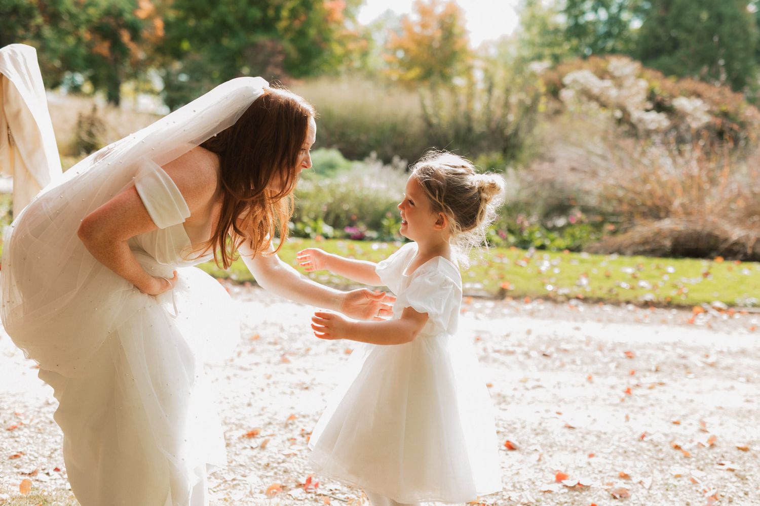 Bride in white wedding gown and veil dancing with flower girl in white dress outdoors at fall wedding.