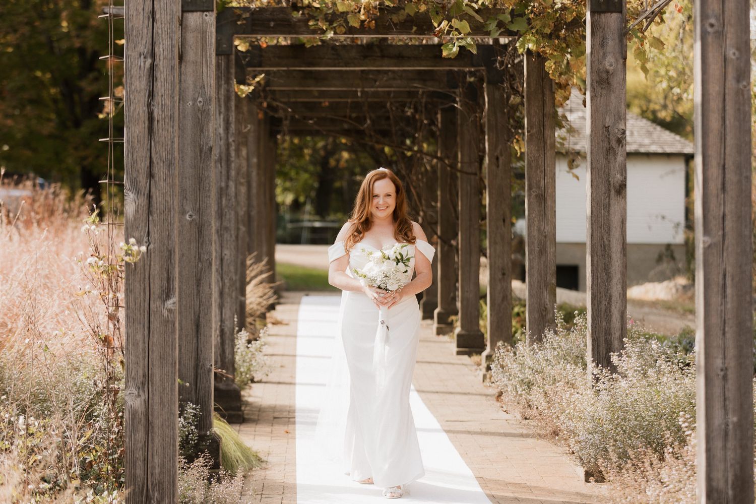 Bride in white gown holding bouquet walks under rustic wooden pergola surrounded by lush greenery on sunny day.