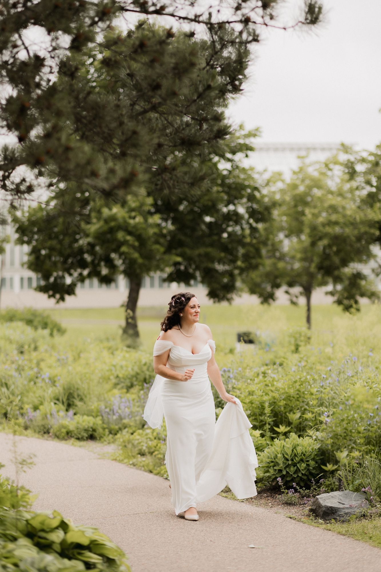 Bride in white off-shoulder gown walks along garden path, holding dress train, surrounded by lush greenery.