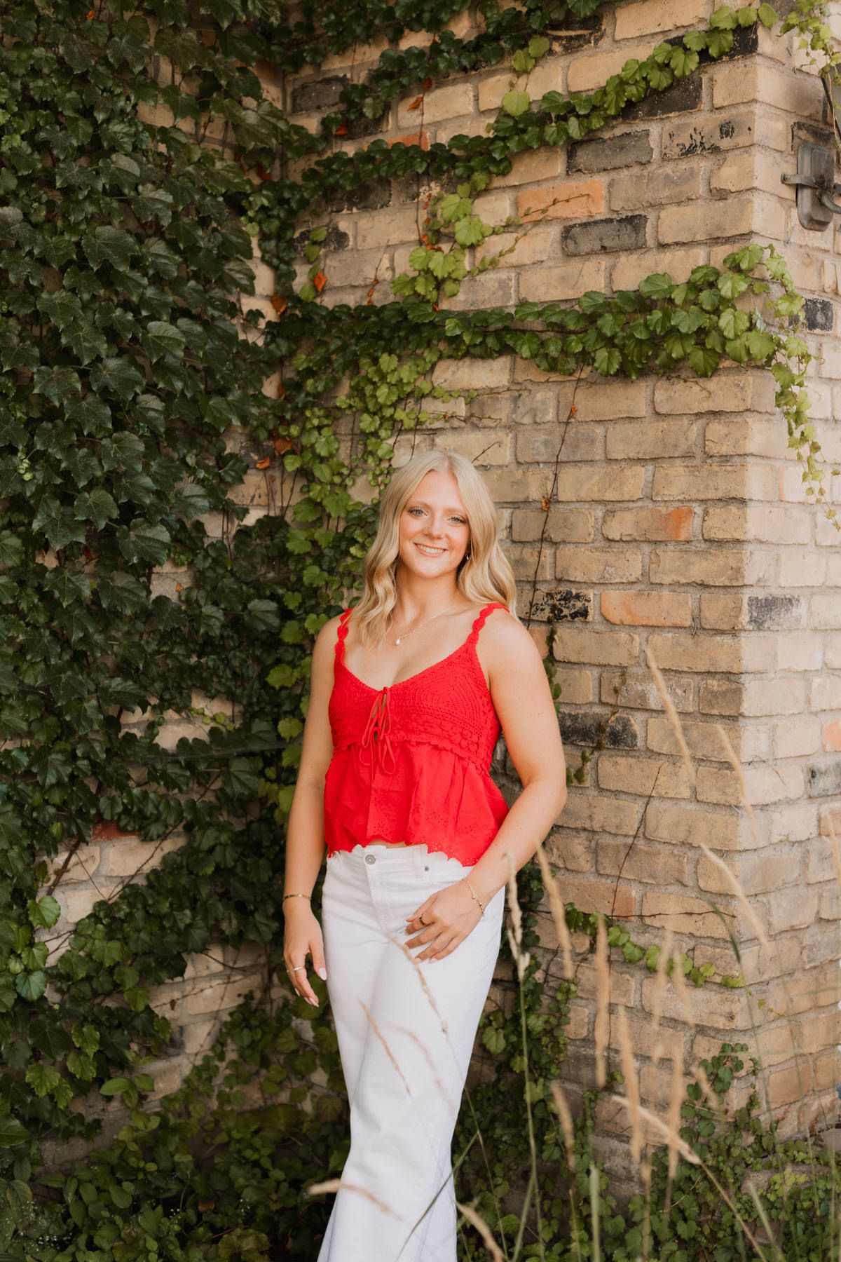 Smiling blonde woman in red top and white jeans leaning against ivy-covered brick wall outdoors.