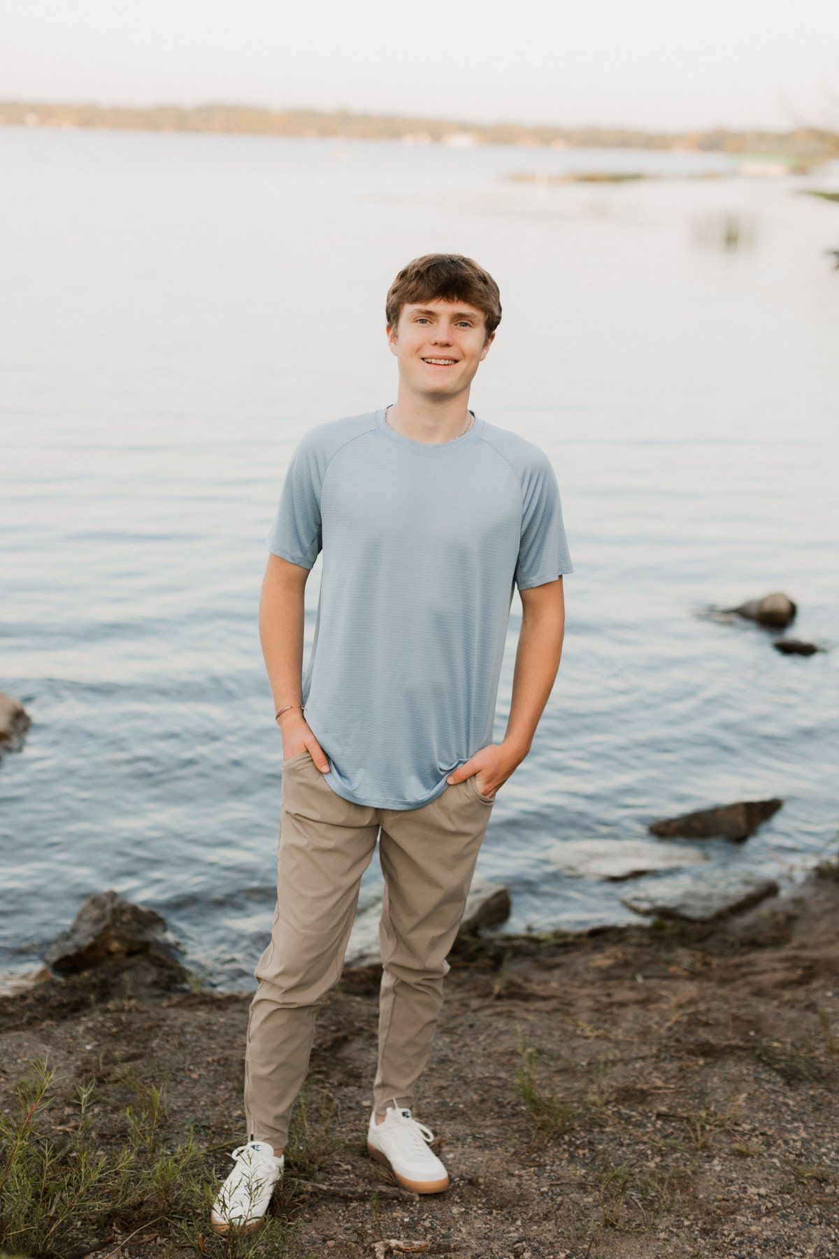 Young man in blue shirt and khaki pants smiling by rocky lakeside shore in natural light.