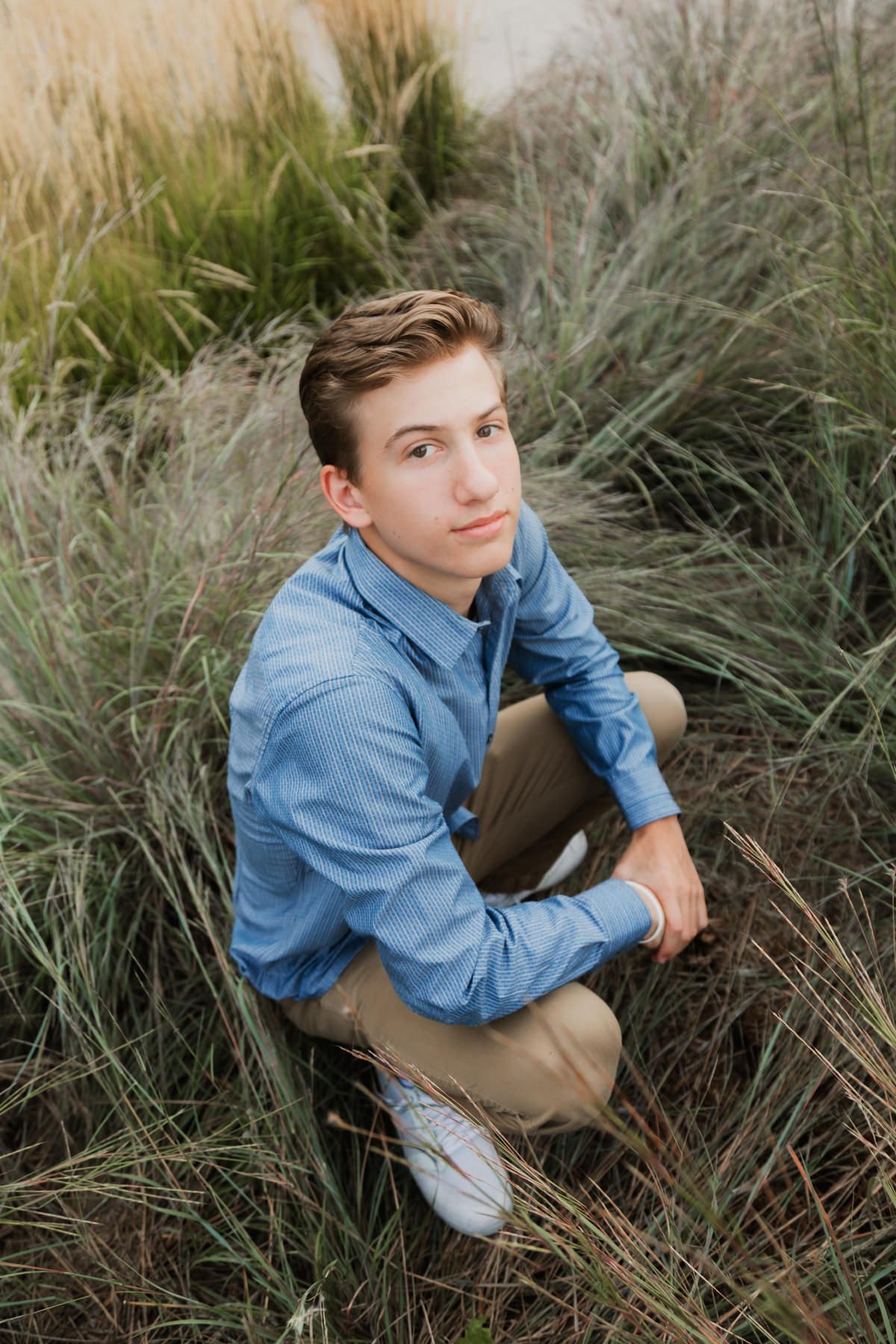 Young man in blue shirt and khaki pants crouching in tall green grass, looking up at camera outdoors.