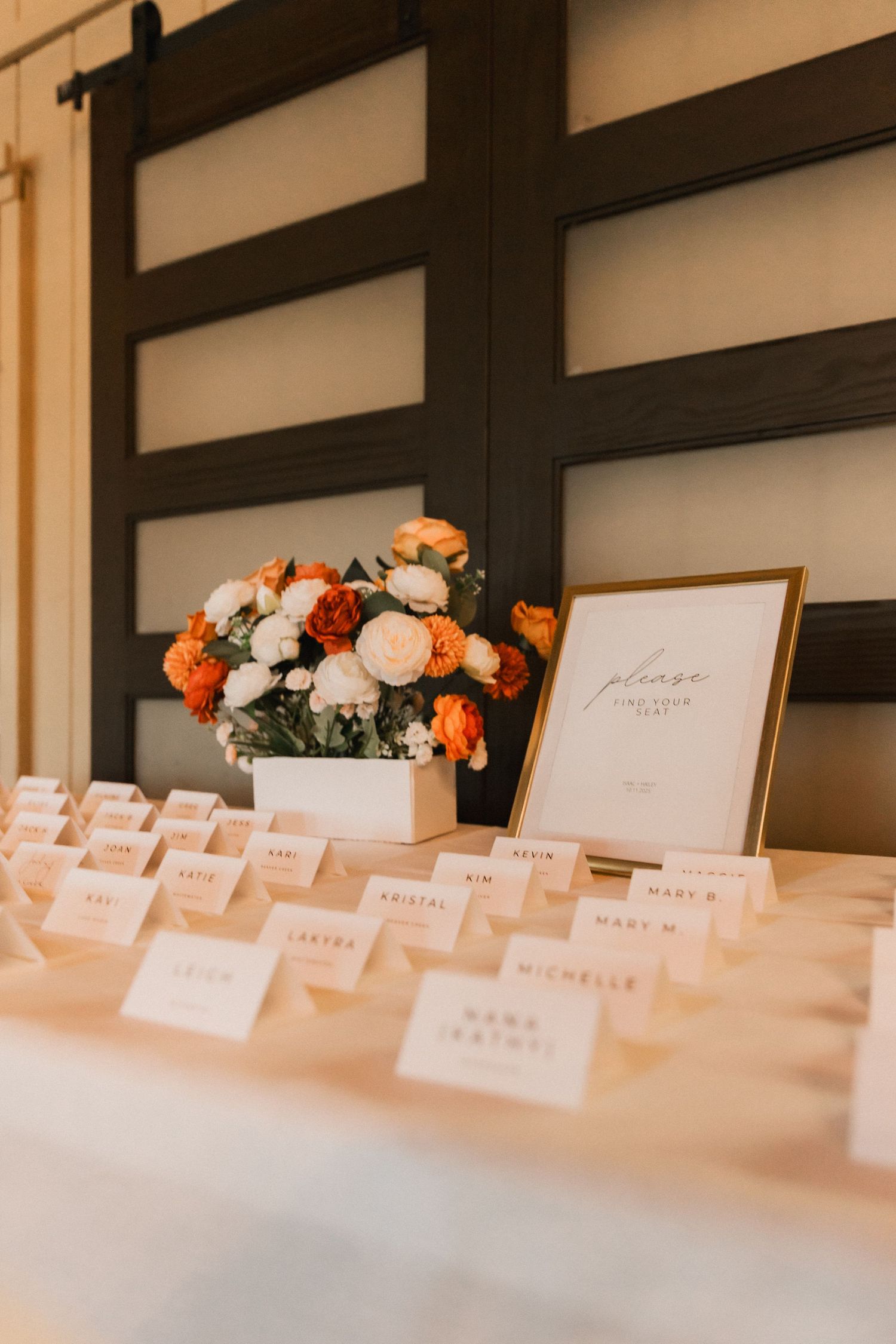 Wedding reception place cards and floral centerpiece with orange and white roses displayed on elegant white table.