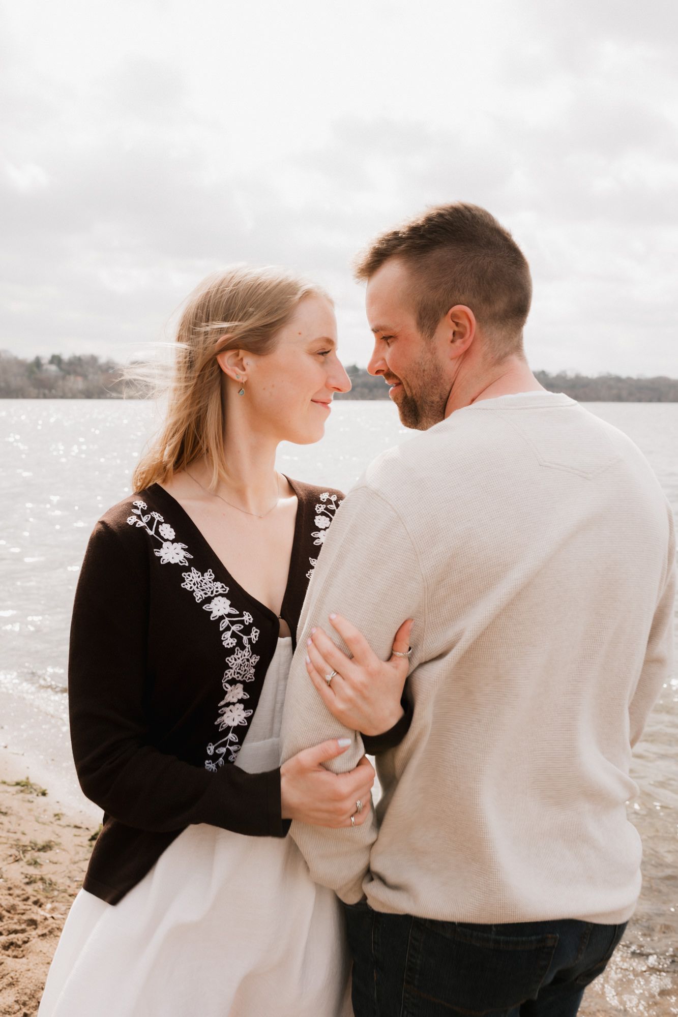 Couple embracing and smiling at each other on a sandy beach by a lake on a cloudy day.