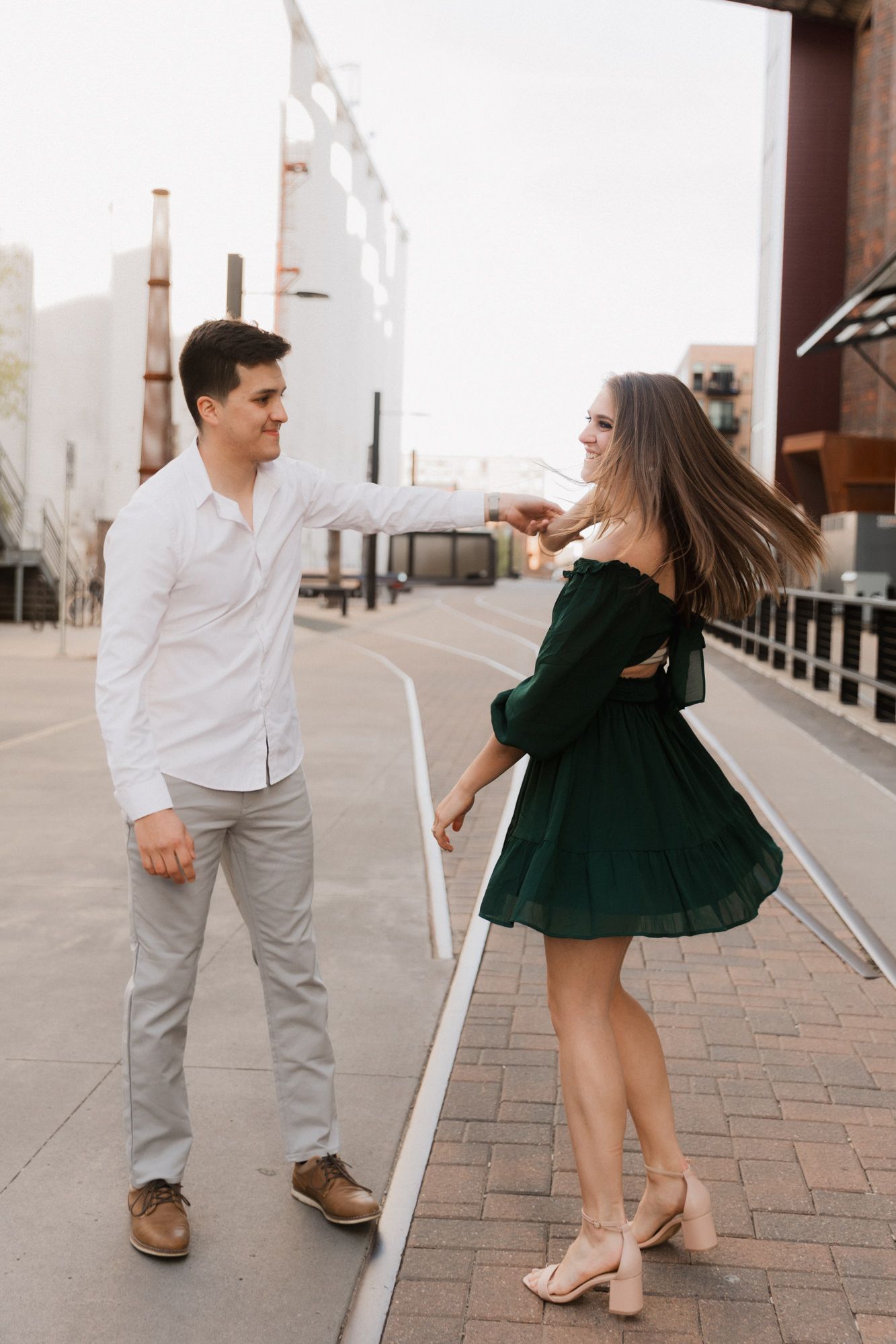A couple dances outdoors, man in white shirt and gray pants, woman in green off-shoulder dress on urban street.